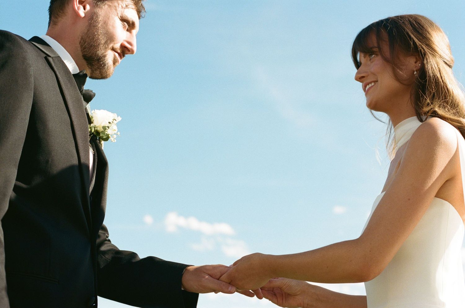 Bride and groom holding hands facing each other under bright blue sky during outdoor wedding ceremony.