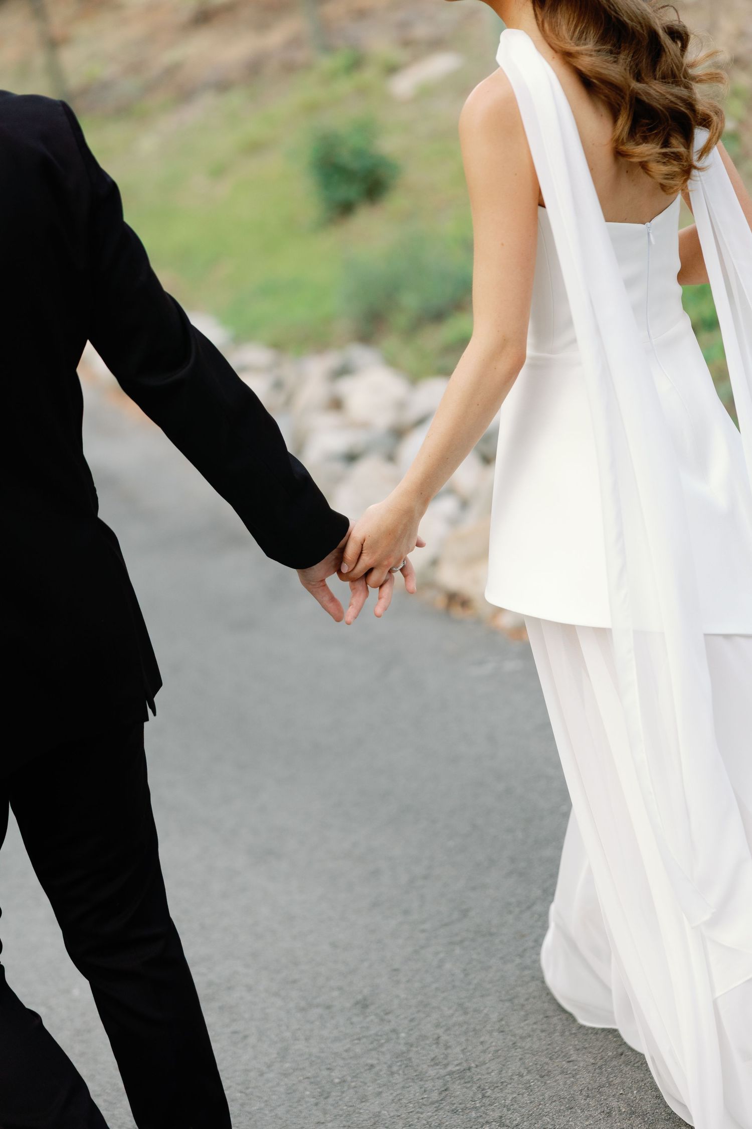 Close-up of bride in white layered gown and groom in black suit holding hands while walking on a road.