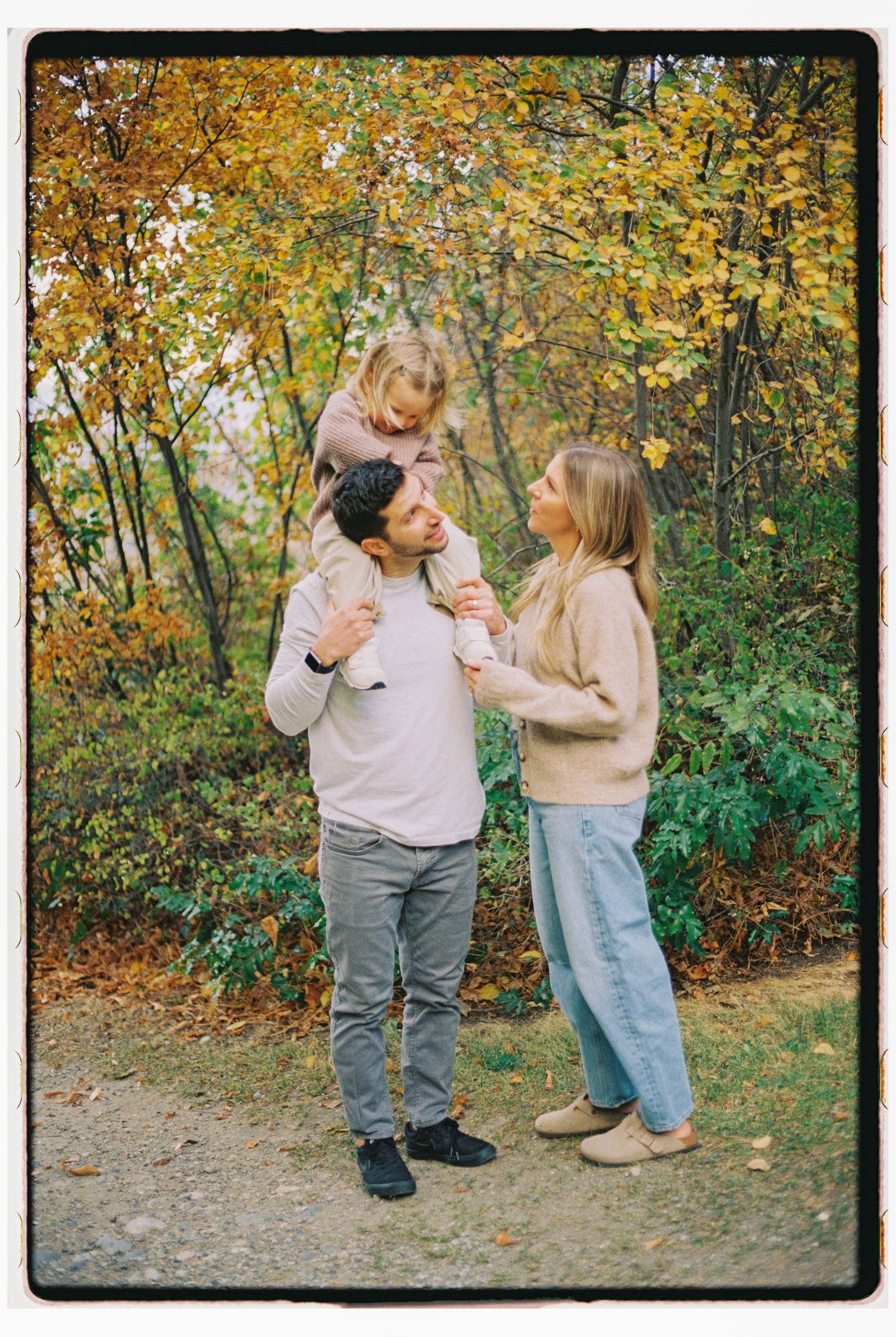 Family of three enjoying autumn park, father carrying toddler on shoulders while mother smiles beside them.