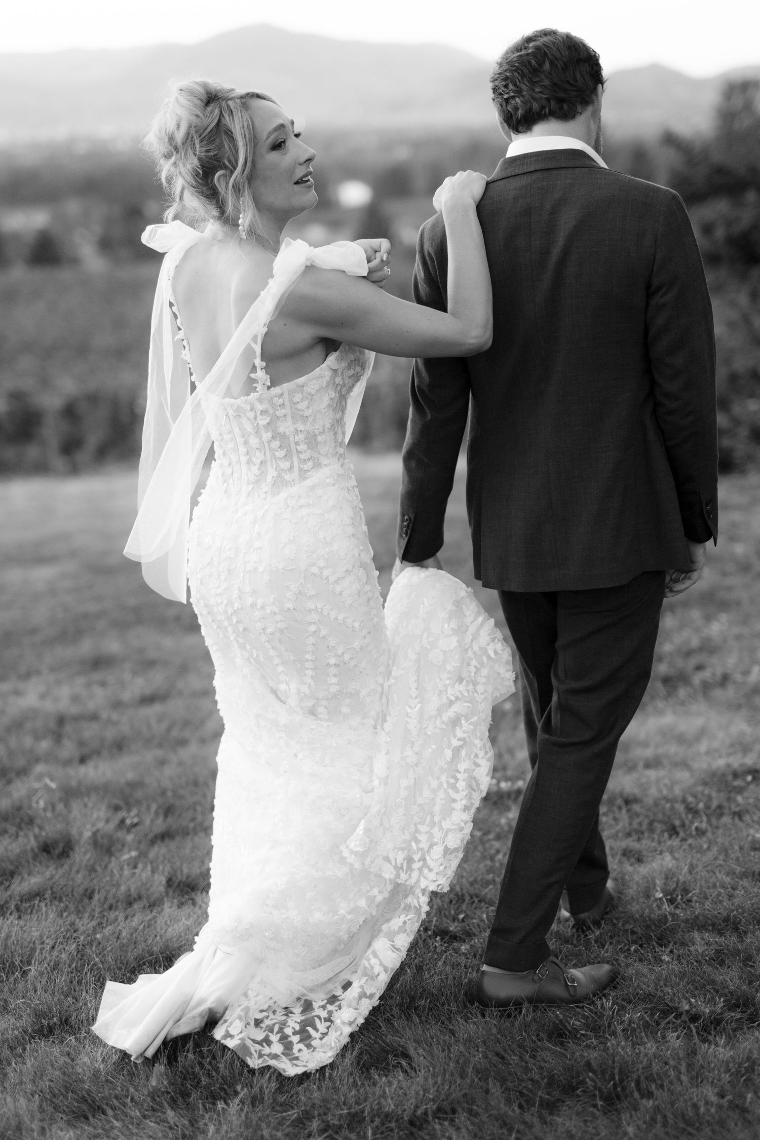 Black and white photo of smiling bride in lace gown walking with groom through scenic outdoor venue.