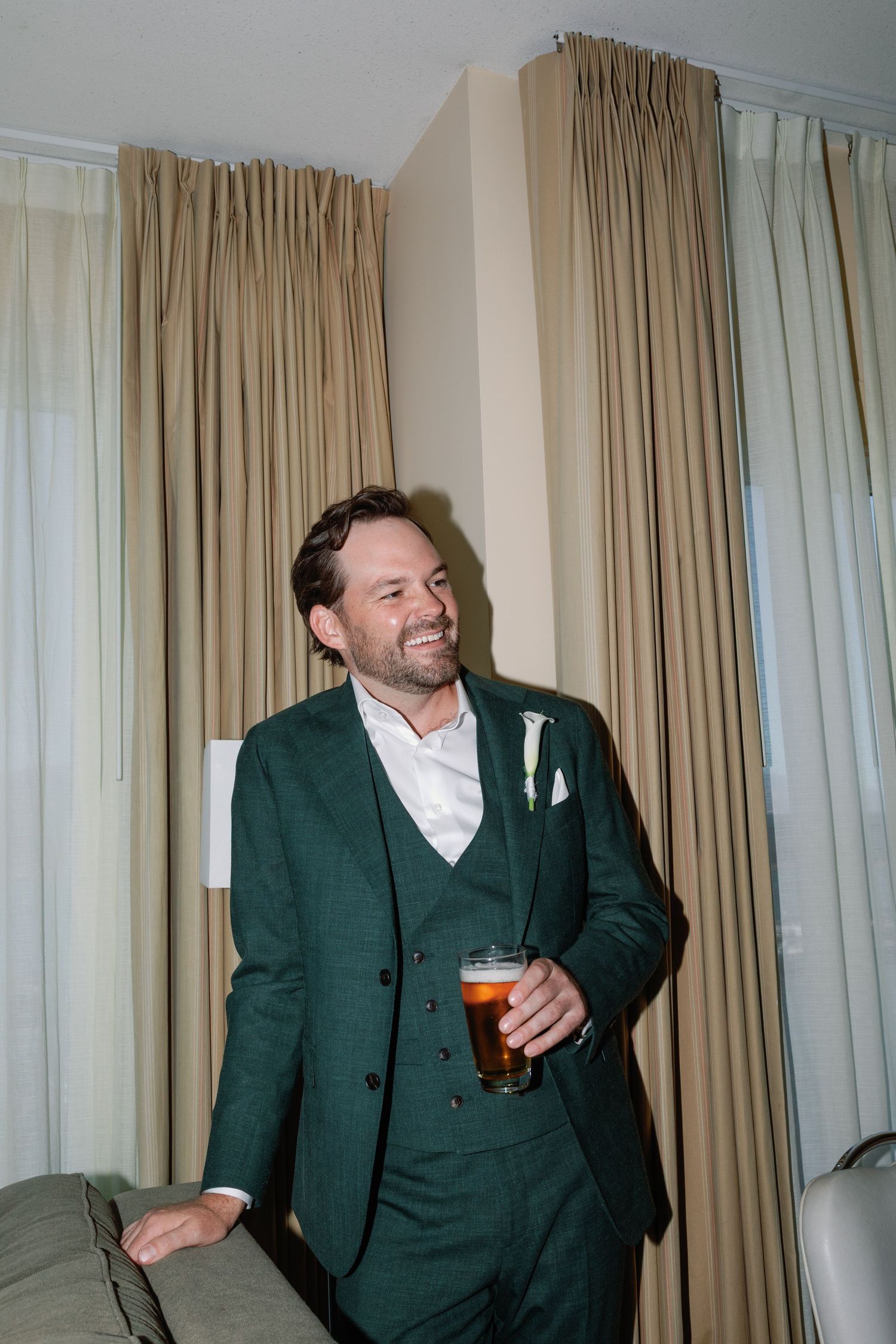 Smiling groom in emerald green three-piece suit holds cocktail glass while standing near beige curtains indoors.