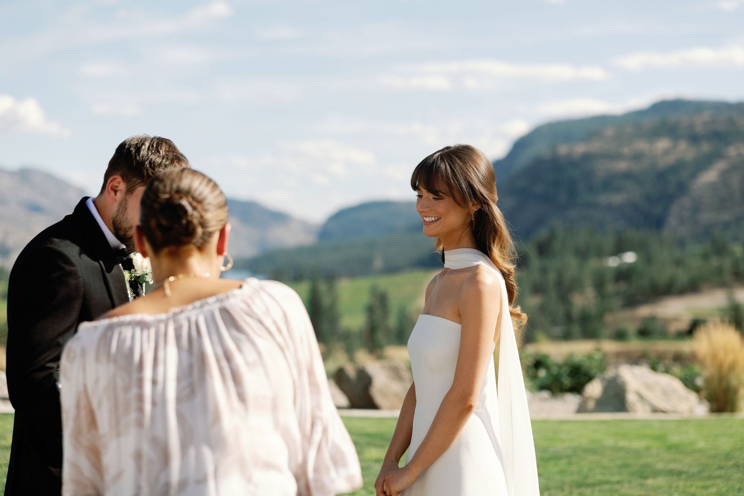 Bride smiling joyfully during outdoor wedding ceremony with officiant and groom against mountain backdrop.