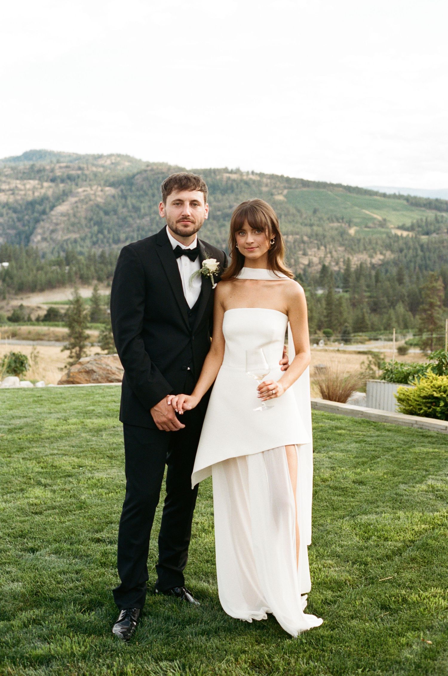 Bride in strapless white gown and groom in black tuxedo pose together with mountain backdrop.