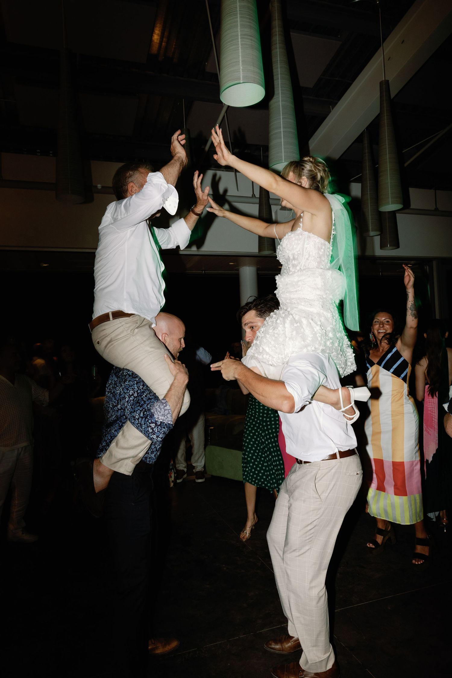 Bride and groom lifted by guests on shoulders during energetic wedding reception dance.