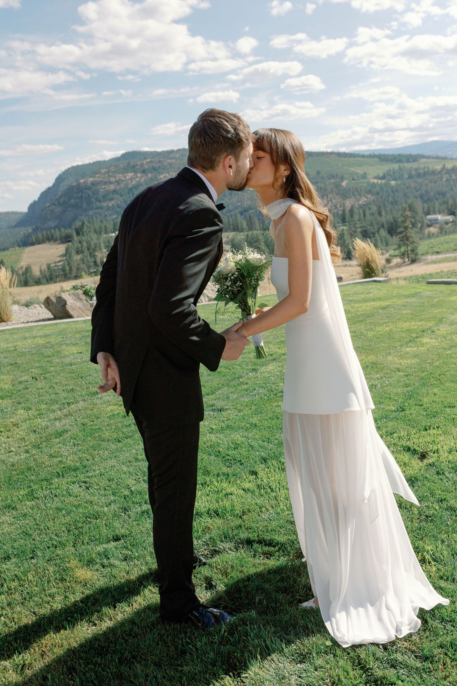 Bride and groom sharing a kiss on a green lawn with scenic mountain backdrop.