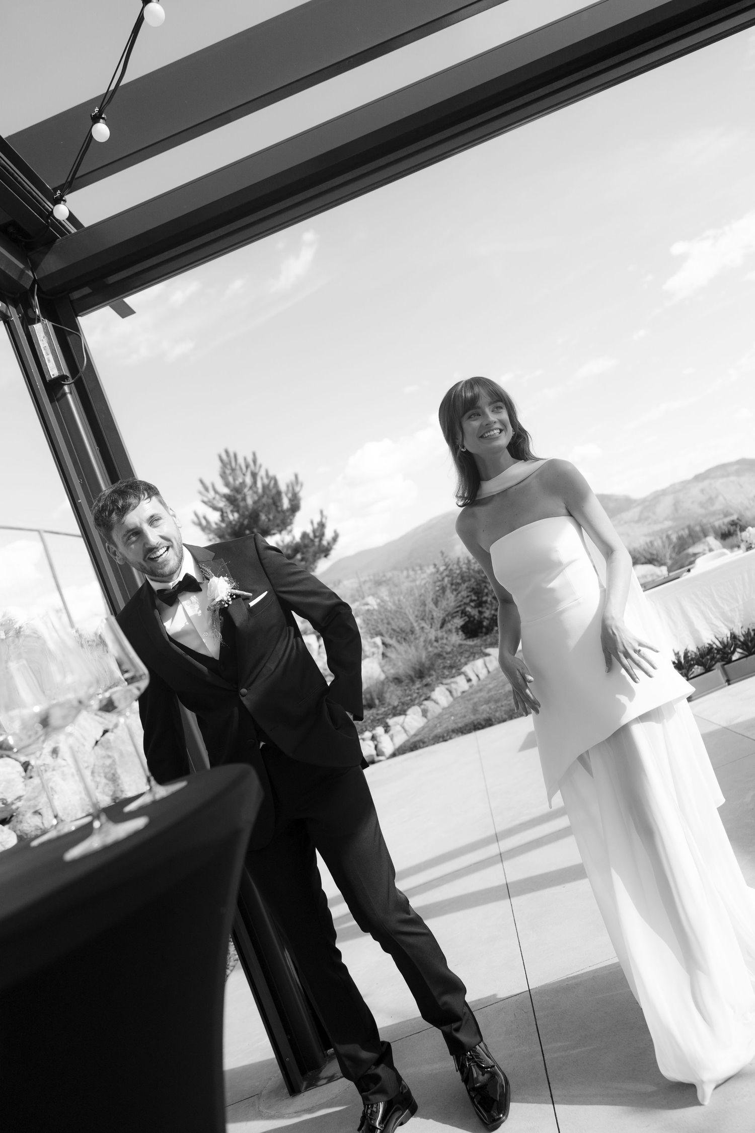 Black and white photo of smiling bride and groom standing by glass doors at reception.