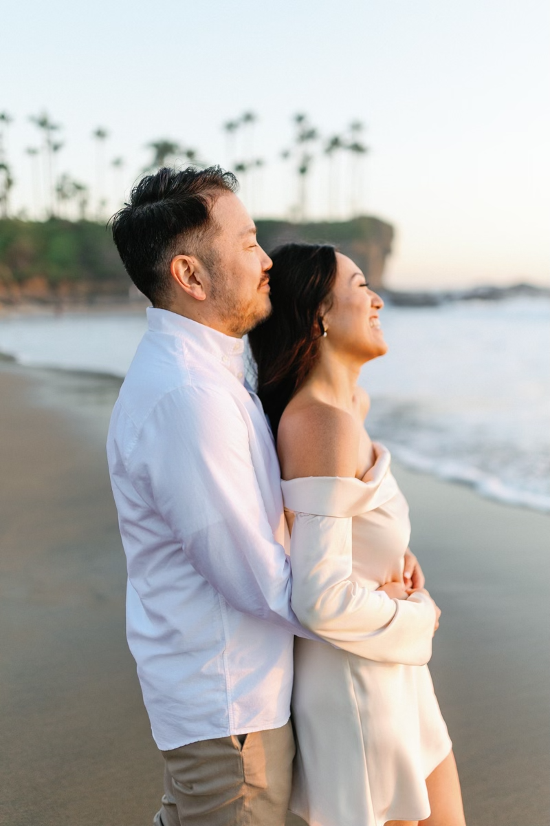Man embracing woman from behind on a beach at sunset, both smiling with palm trees in background.