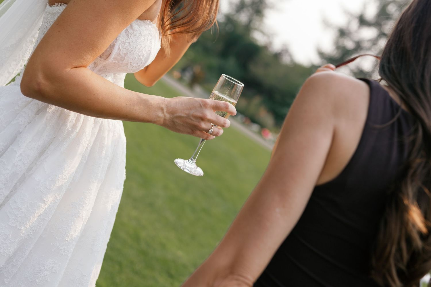Bride in white dress holding champagne flute chatting with guest in black dress at outdoor wedding reception.
