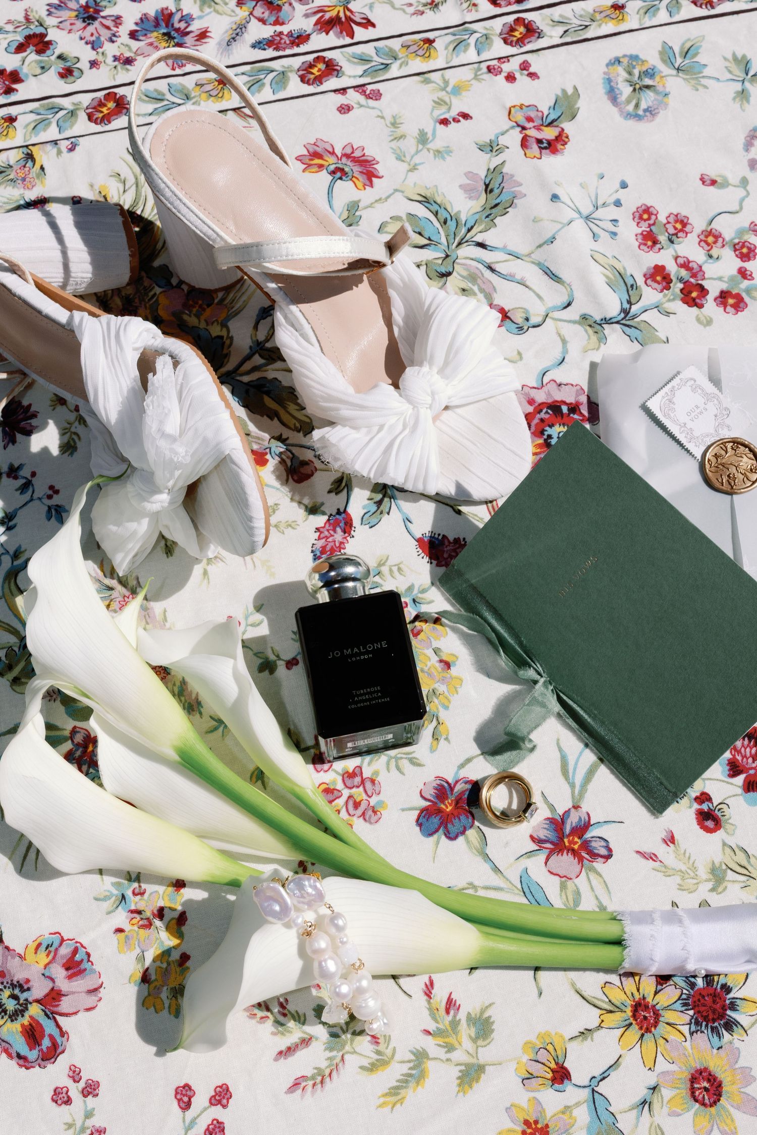 Elegant wedding flat lay with white heels, calla lily bouquet, perfume, green journal, and pearl bracelet on floral fabric.