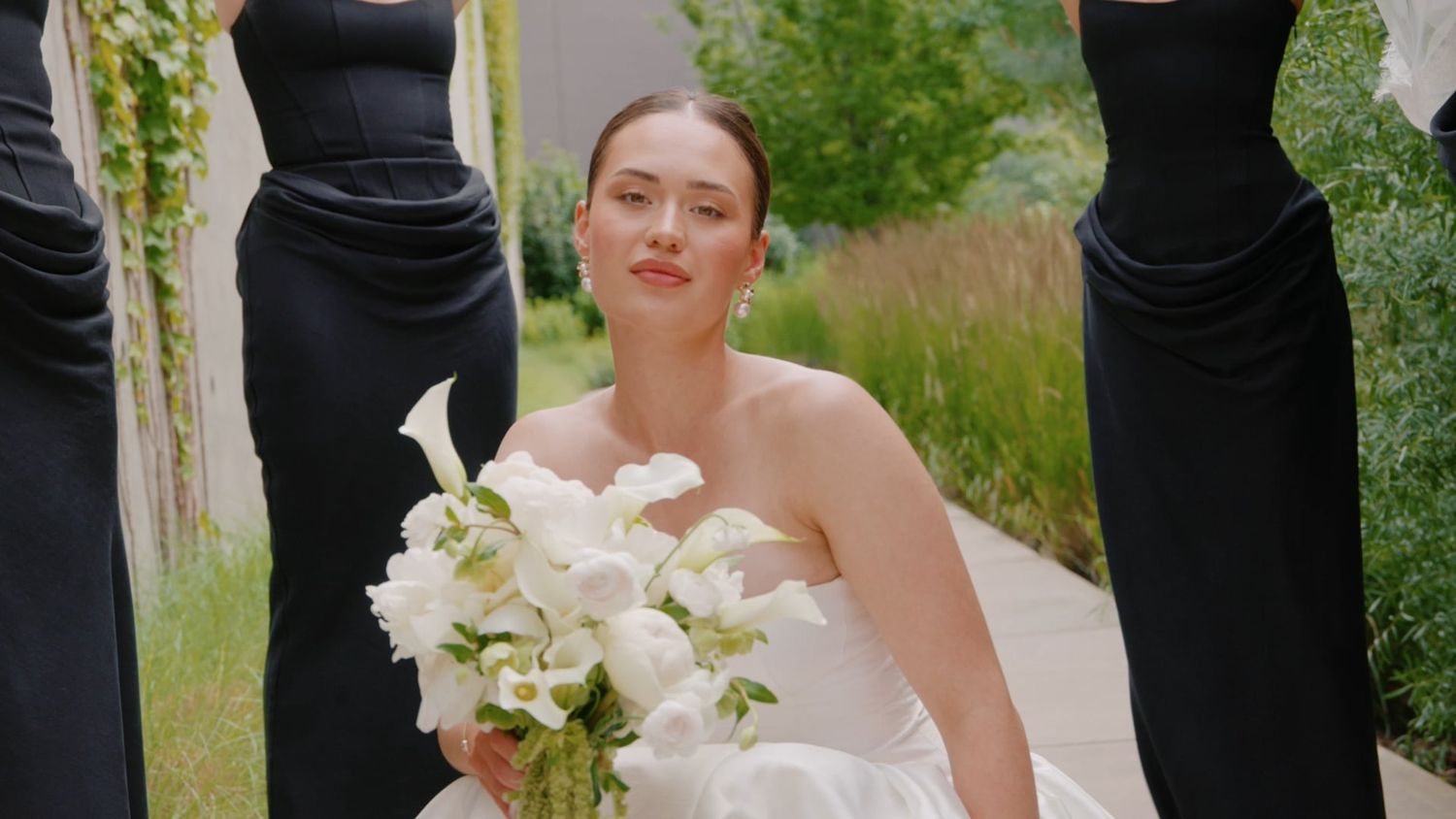 Bride in white gown holds white calla lily bouquet, flanked by bridesmaids in black dresses, outdoors.