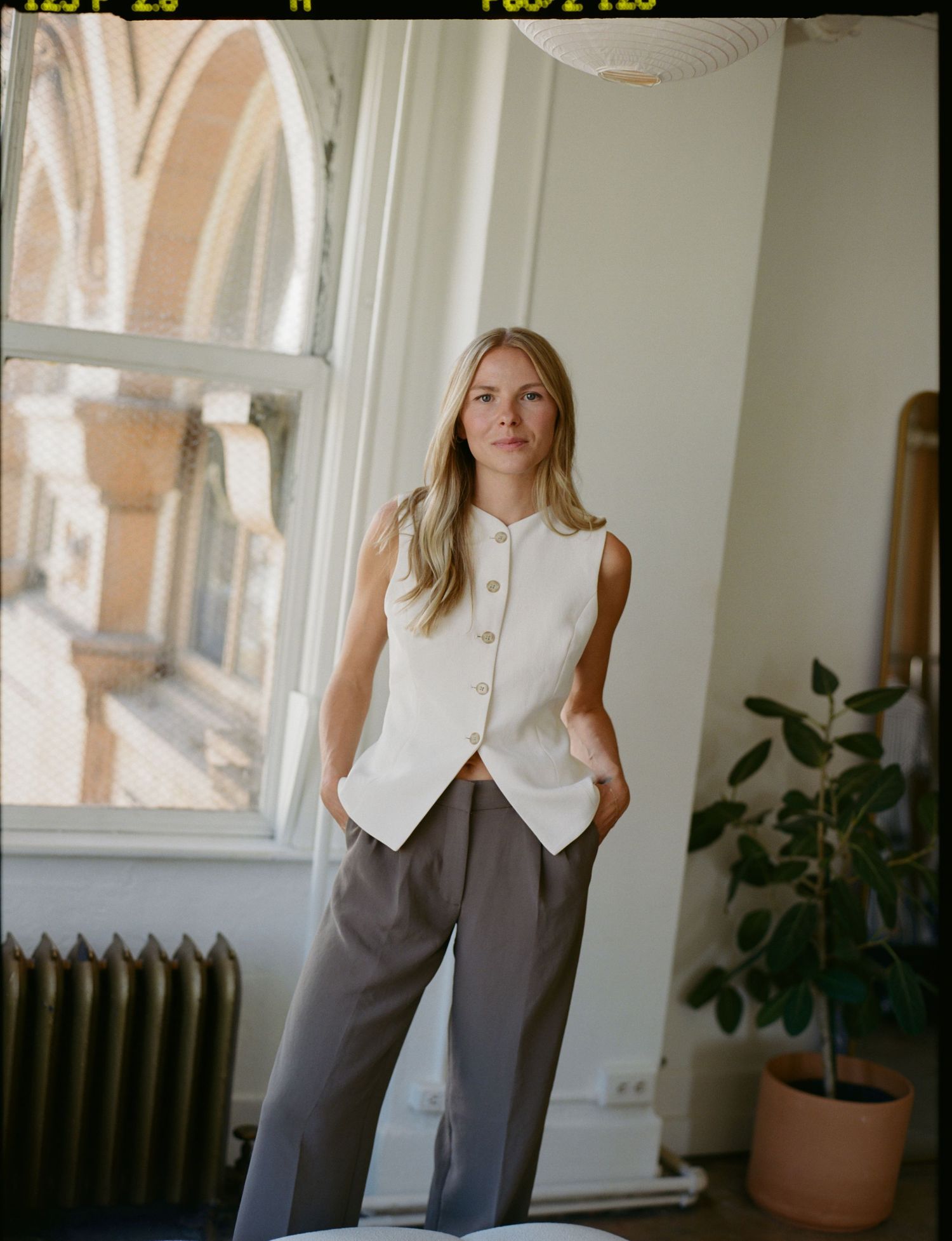 Woman in white sleeveless vest and grey trousers standing by a window in a bright room with a plant.