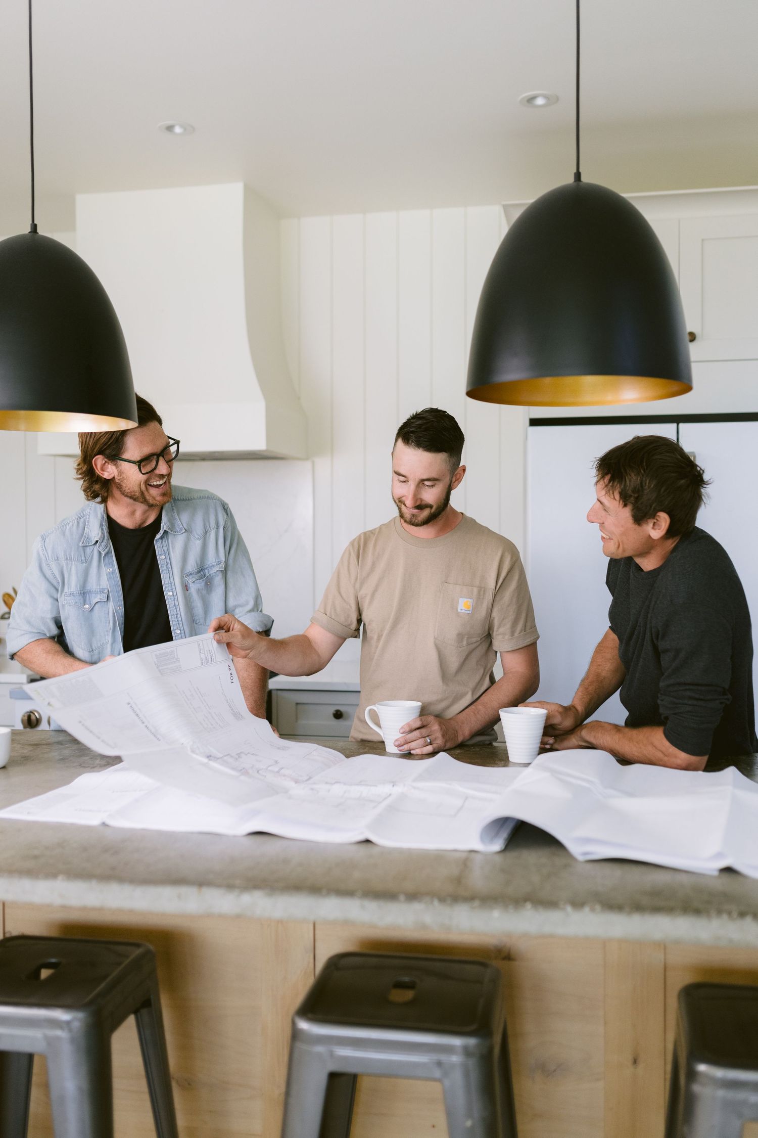 Three men reviewing architectural blueprints at a kitchen island with coffee mugs under black pendant lights.