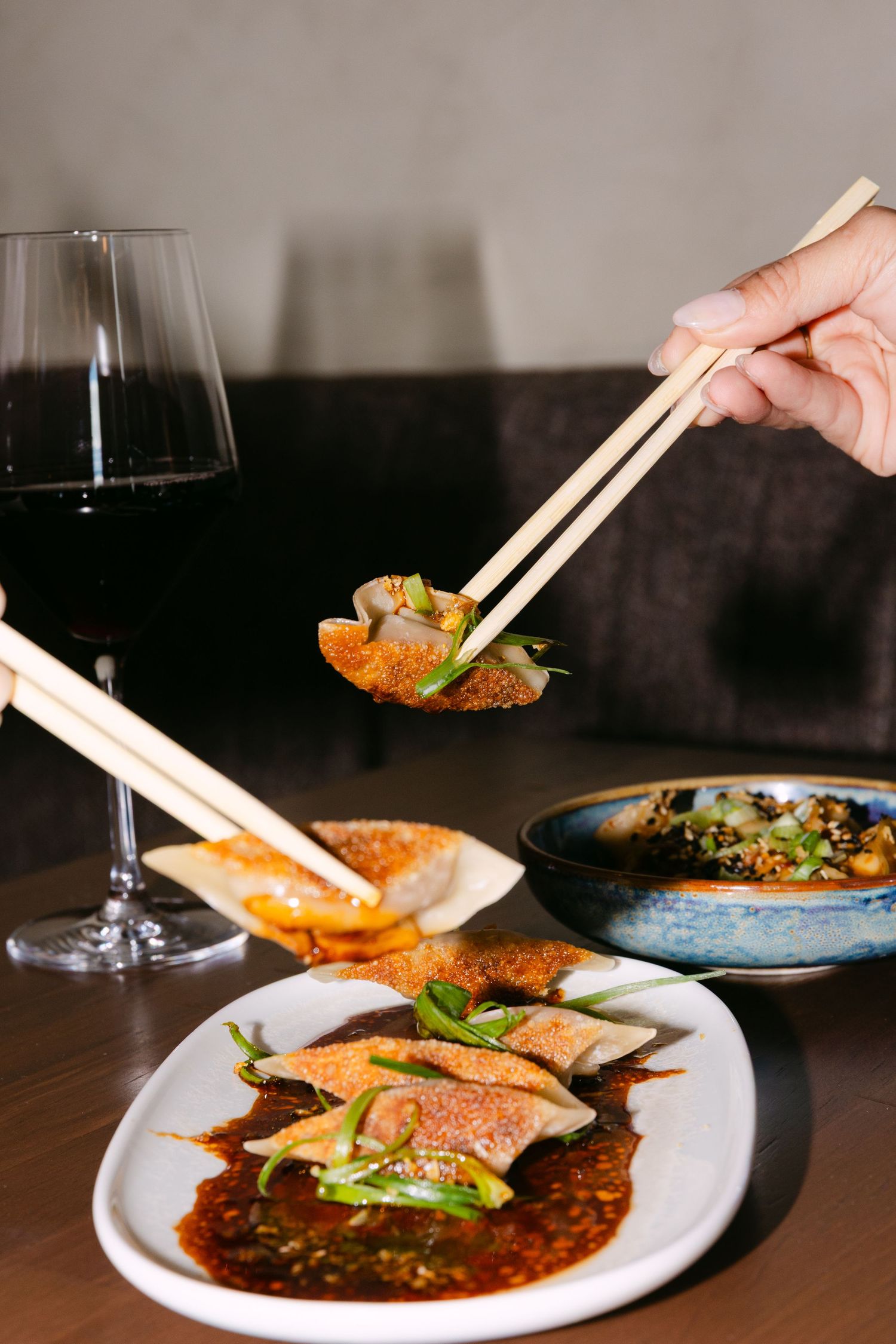 Hands using chopsticks to pick up a dumpling from a plate of spicy sauce dumplings, served with red wine.