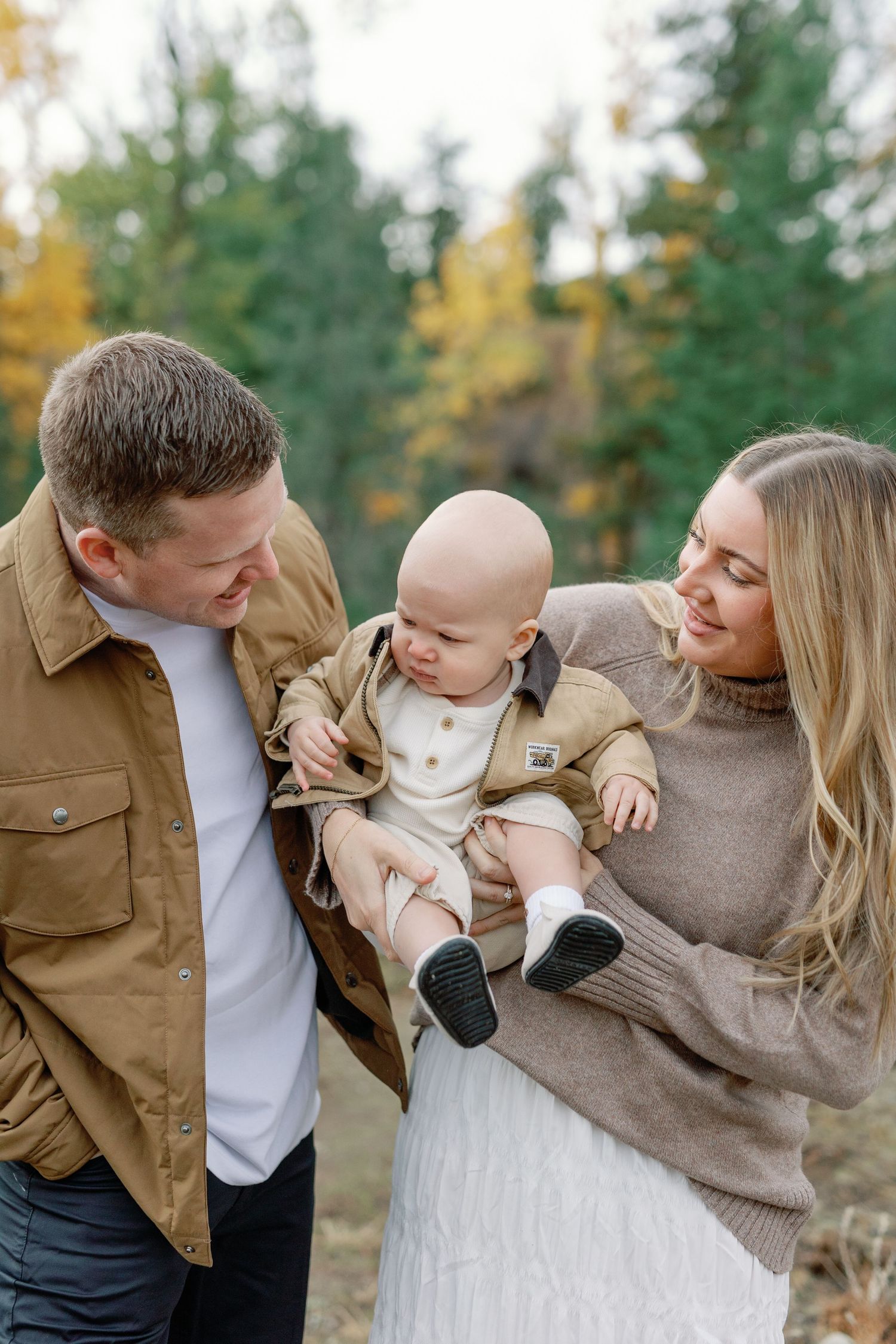 A young couple smiles at their baby boy outdoors in an autumn forest setting, dressed in warm neutral tones.