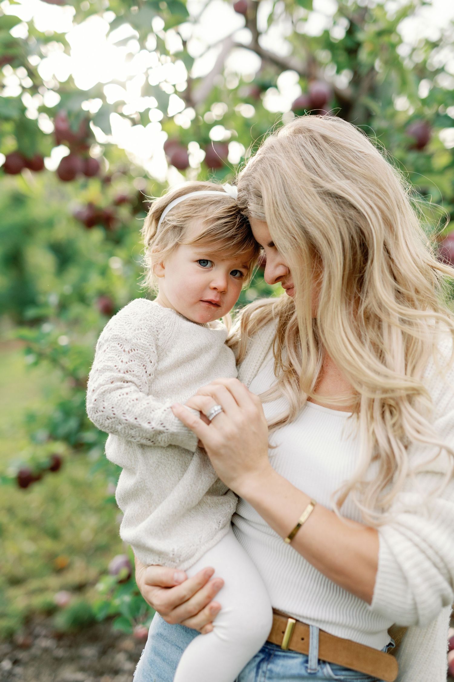 A blonde mother tenderly holds her toddler daughter in an apple orchard, both wearing neutral tones in a warm outdoor setting.