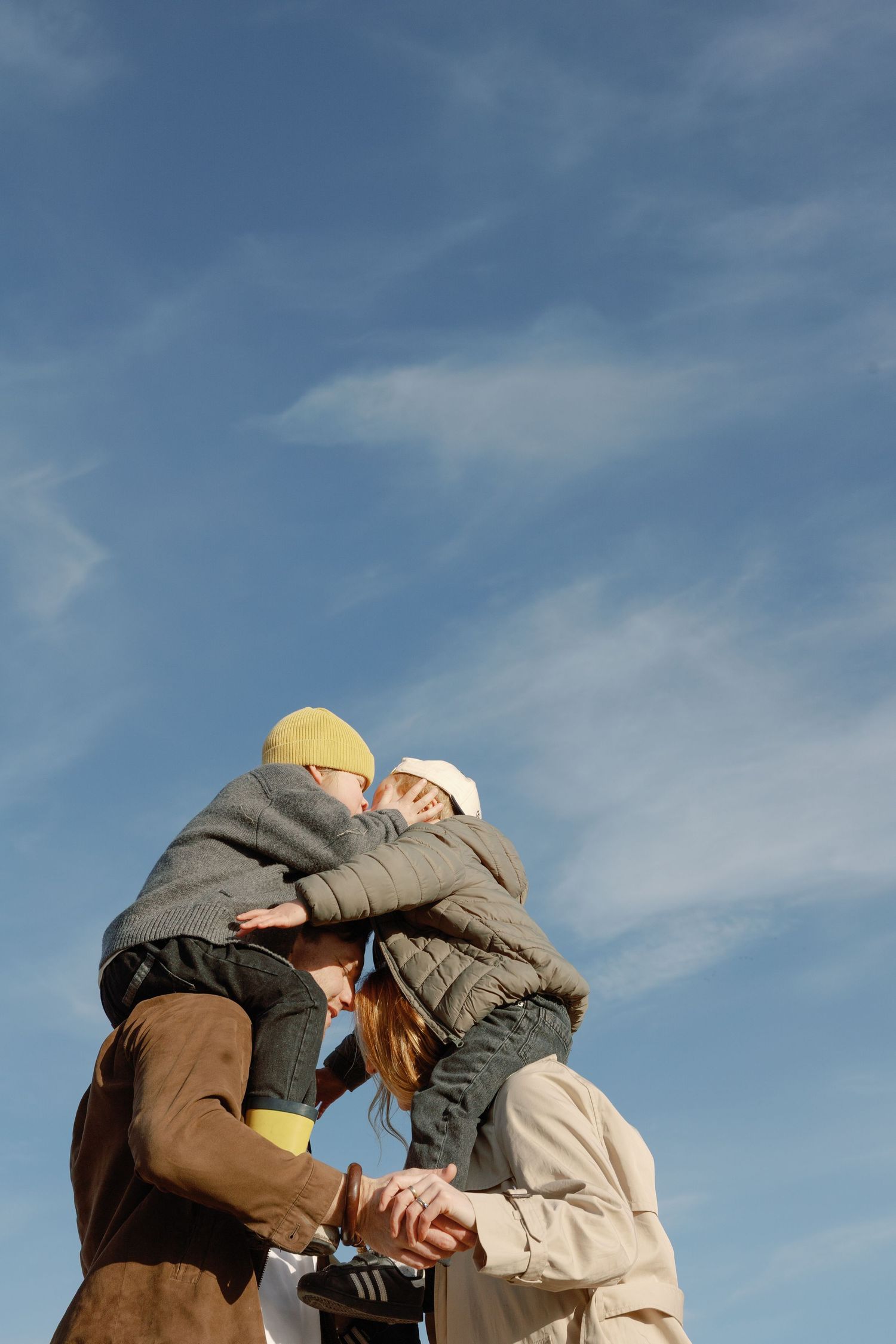 Family embracing outdoors under a blue sky, parents lifting child in warm winter jackets and hats.