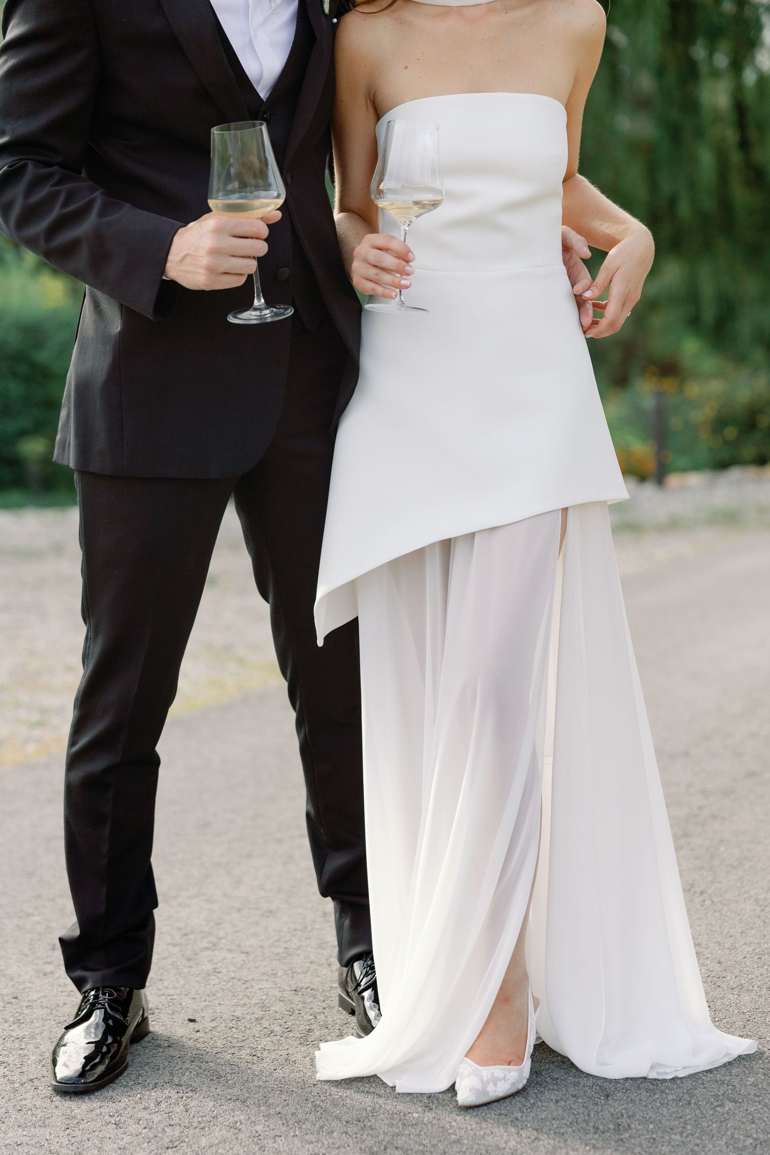 Close-up of bride in strapless white gown and groom in black tuxedo holding champagne glasses on a paved path.