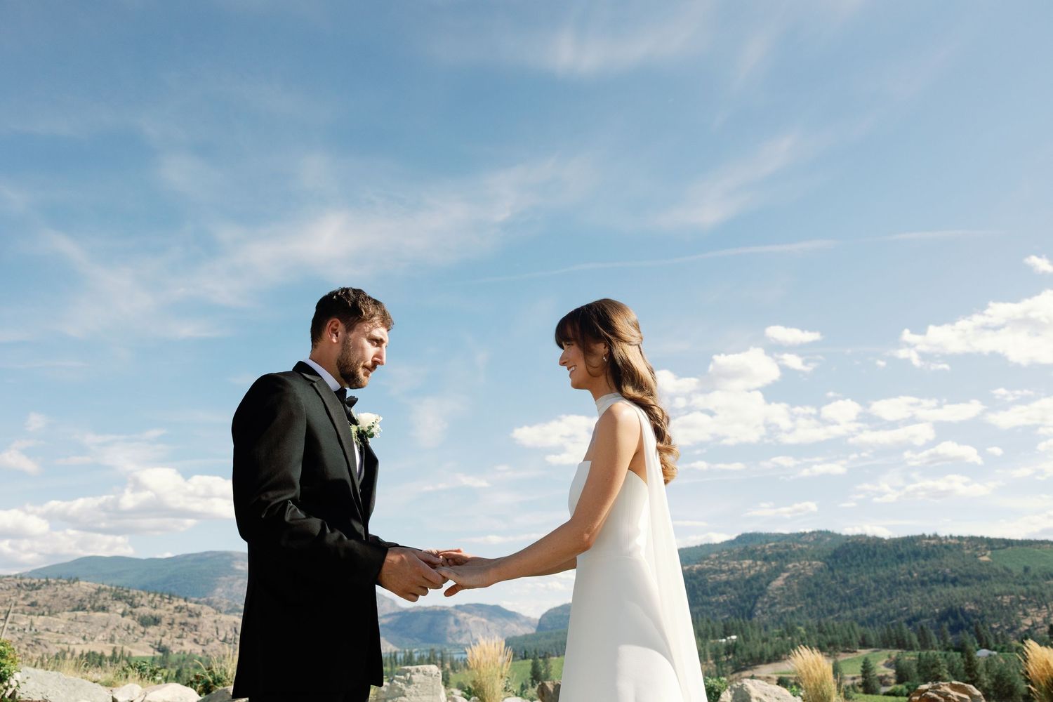 Bride and groom hold hands during outdoor wedding ceremony under blue sky with mountain views.
