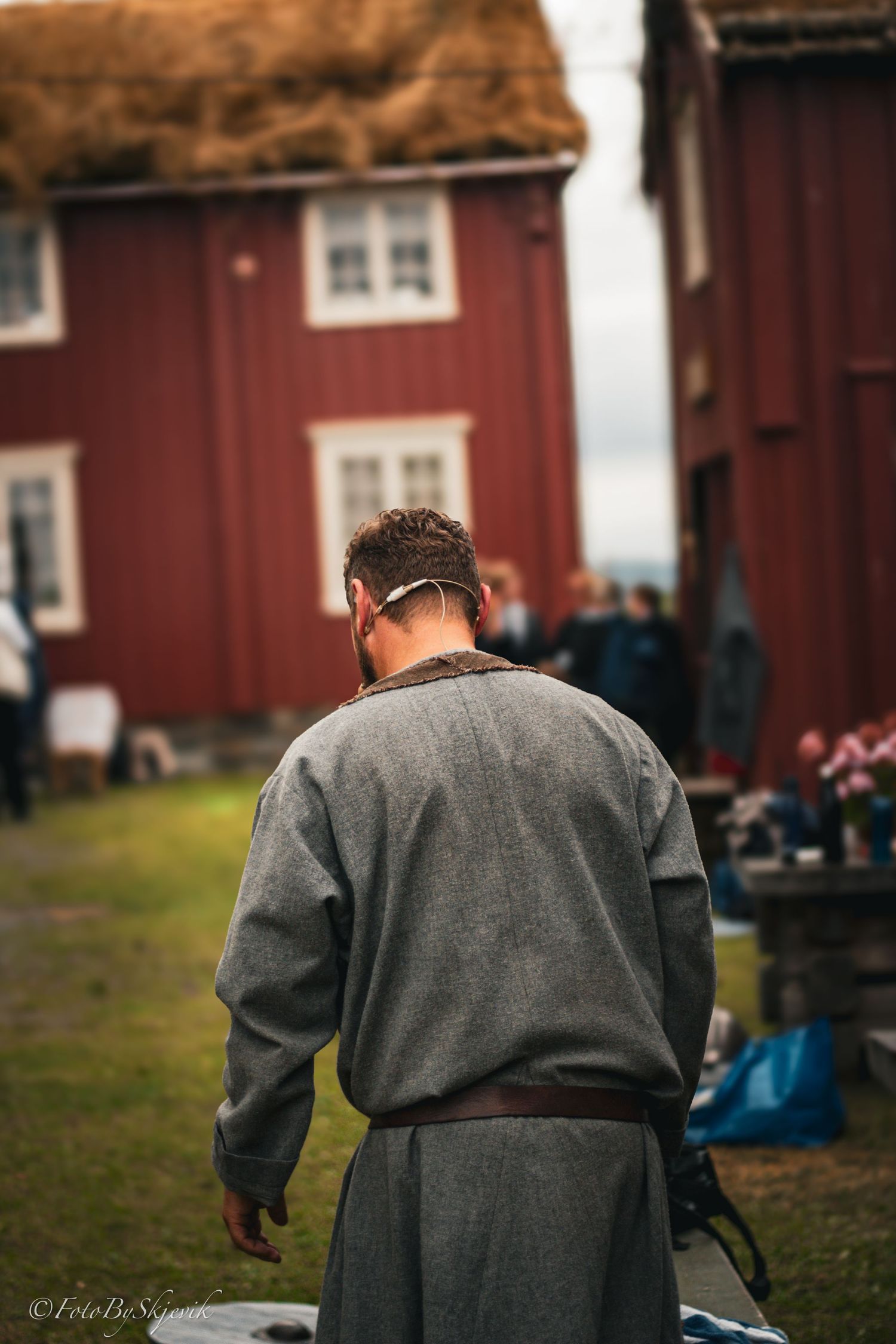 Person wearing a grey medieval-style tunic near red barn buildings.