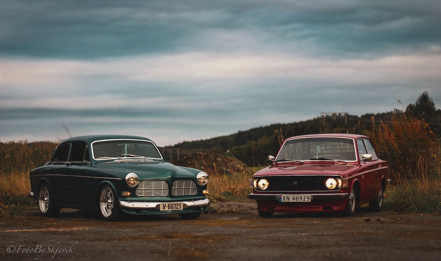 Two vintage cars, a black and burgundy, parked side by side against autumn backdrop.