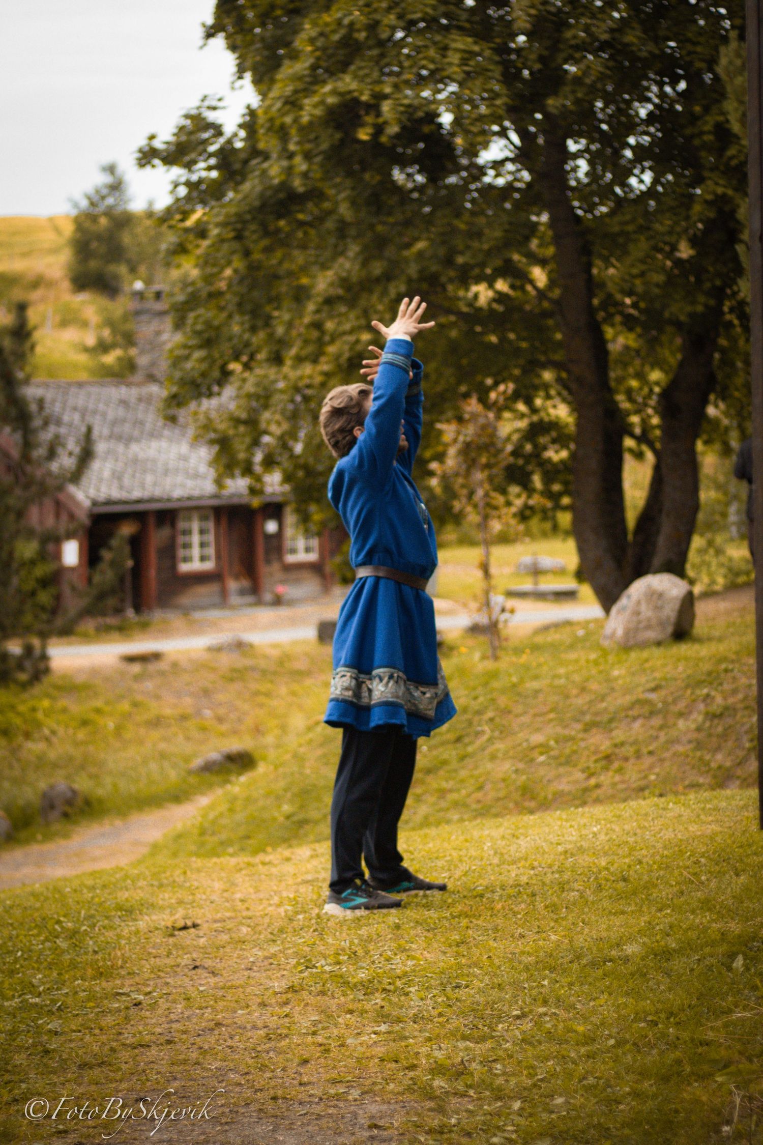 Person in blue traditional costume gestures dramatically on a grassy hillside near red buildings.