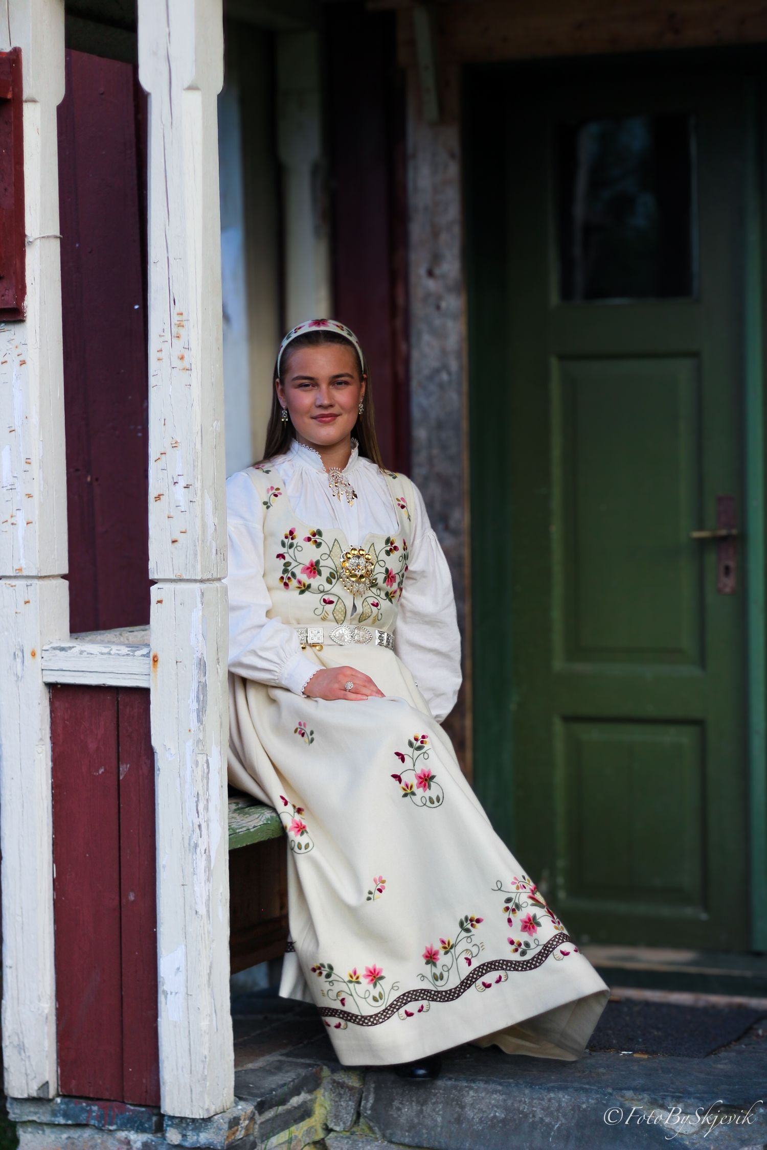 Young woman in traditional Norwegian bunad folk costume with floral embroidery stands by a rustic doorway.