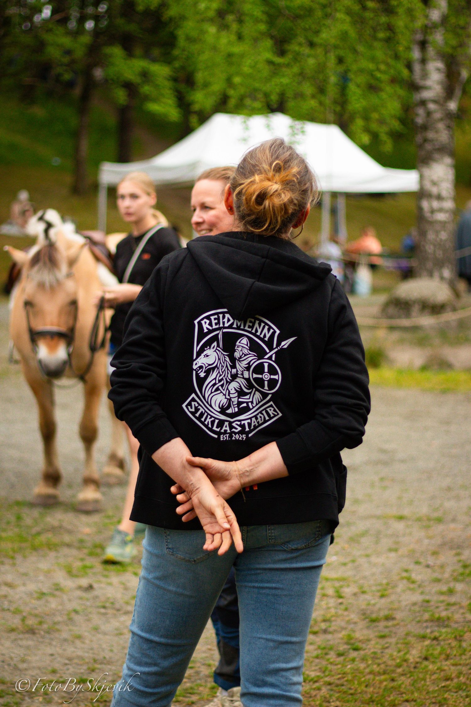 Woman in black Reidkenn Fjordklastabler hoodie stands at outdoor equestrian event with horses and white tent.