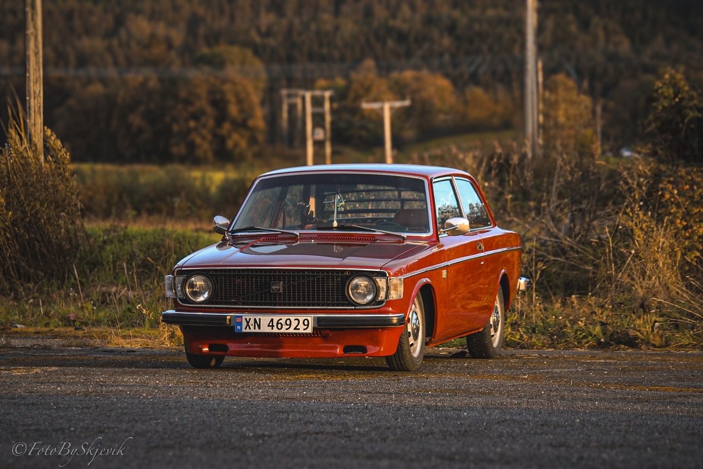 A classic red Volvo coupe parked on a dirt road at sunset with autumn trees in the background.
