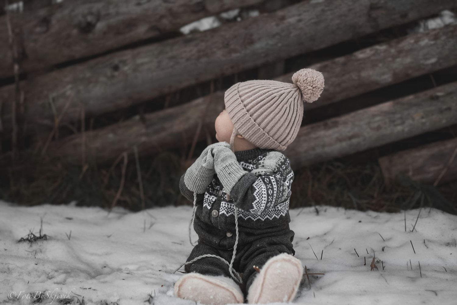 A toddler in a knit pom-pom hat and Nordic sweater sits in snow near rustic wooden logs in winter.