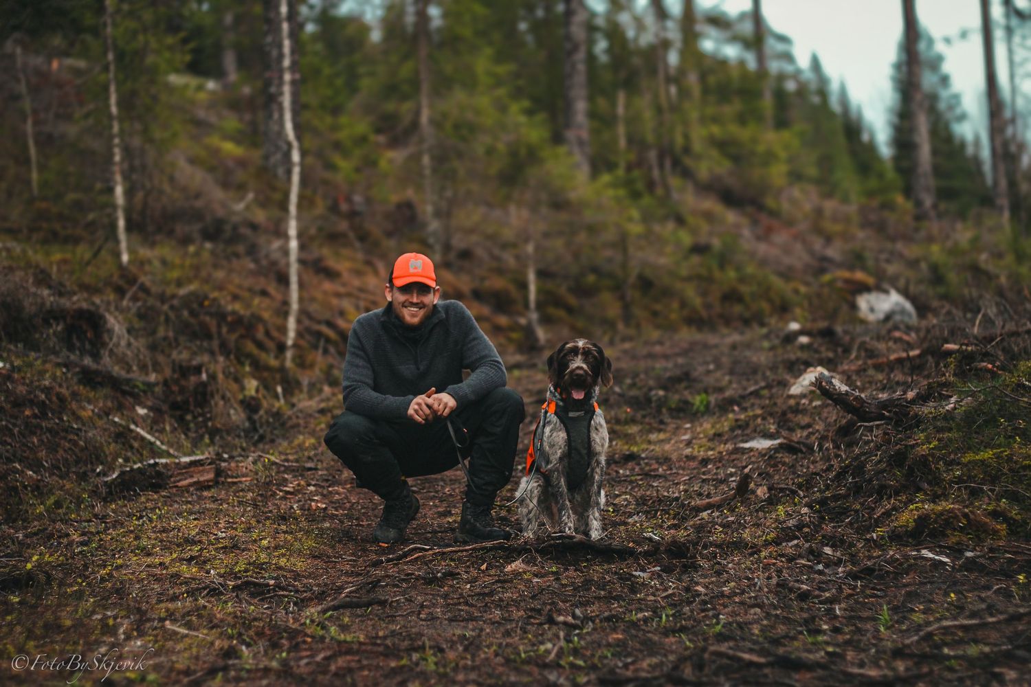 Hunter in orange cap with pointer dog in forest setting.