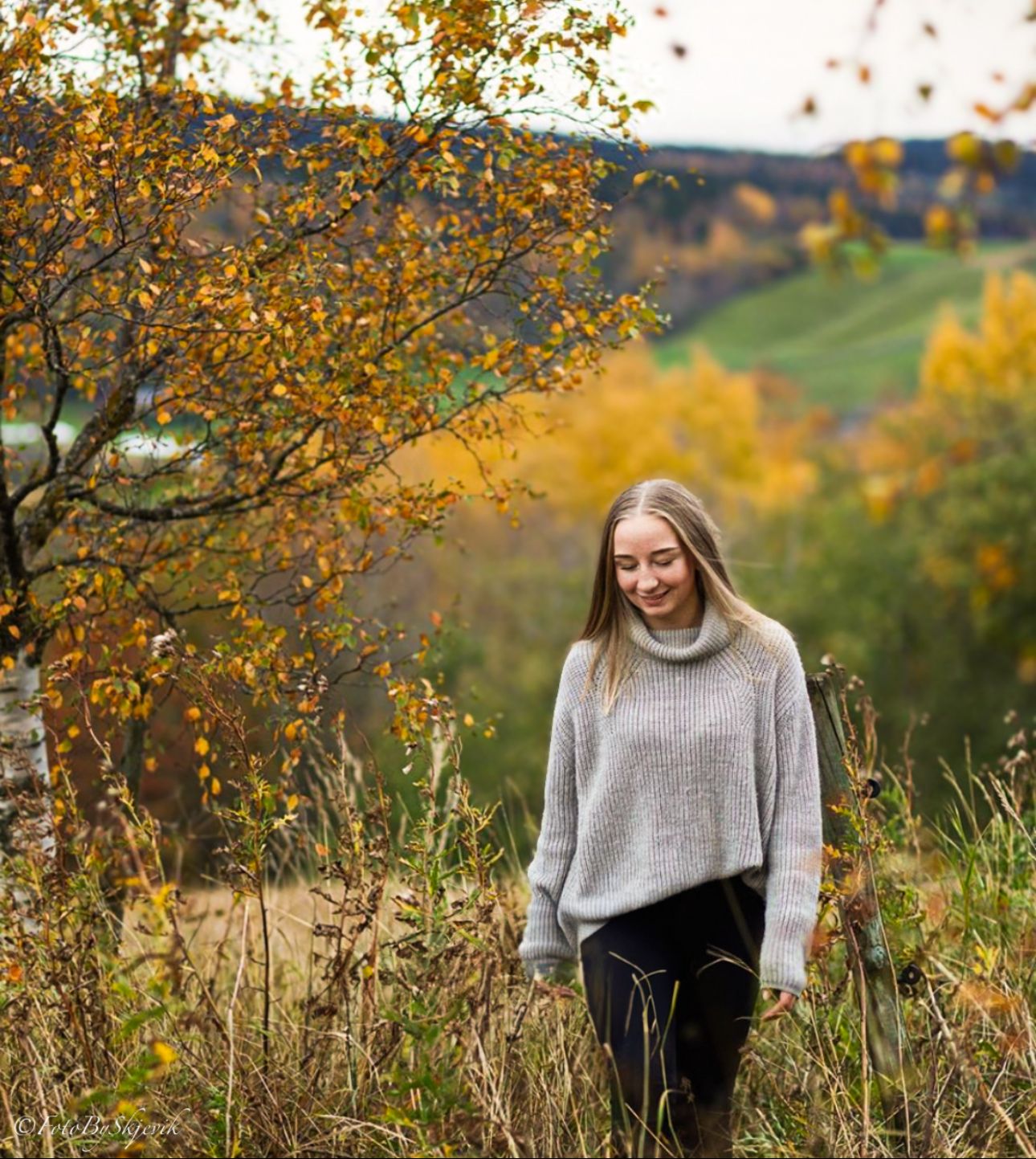 A person in a gray sweater stands among golden autumn foliage on a hillside during fall season.