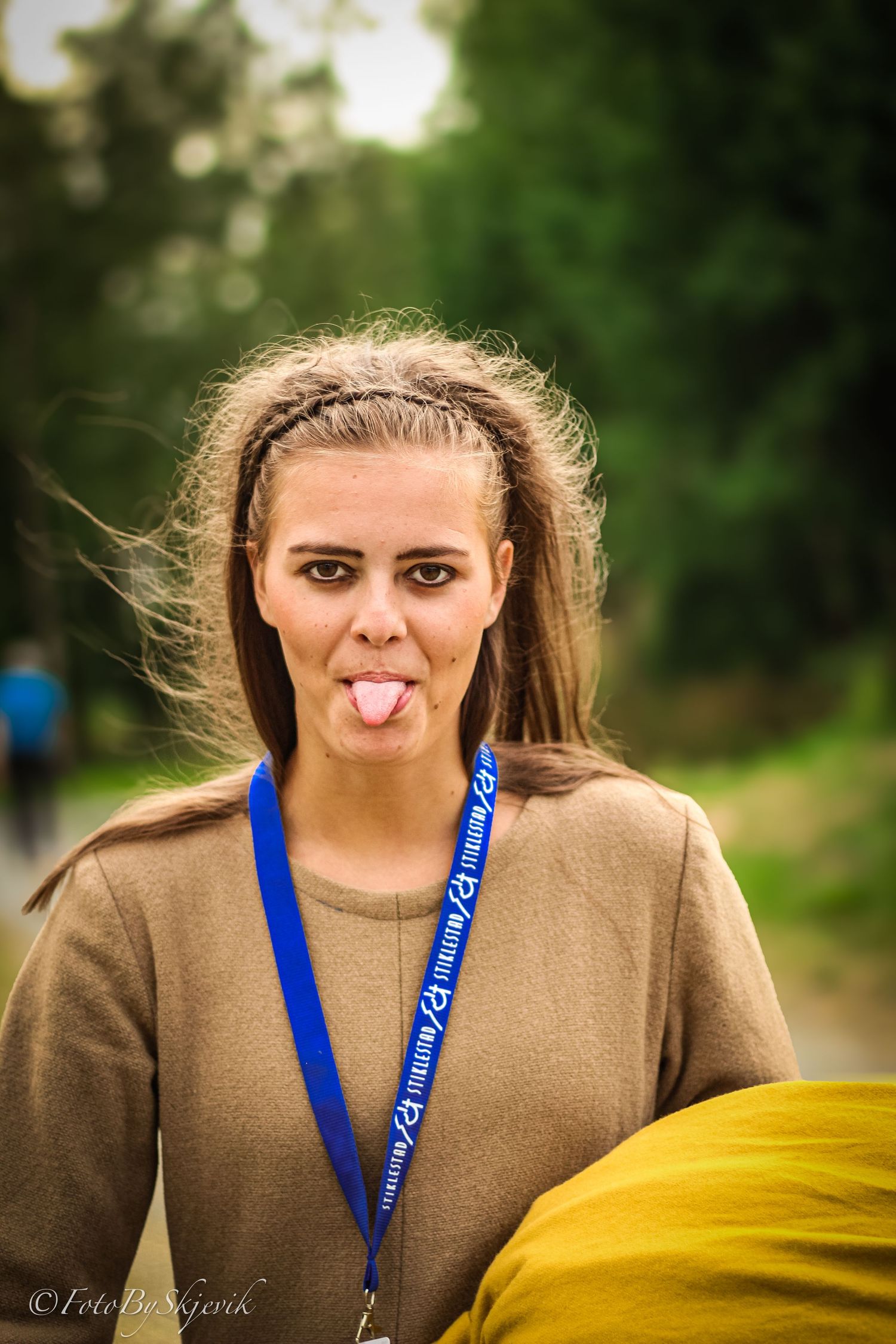 Young woman sticking out her tongue outdoors, wearing a brown sweater and blue lanyard, with trees in the background.