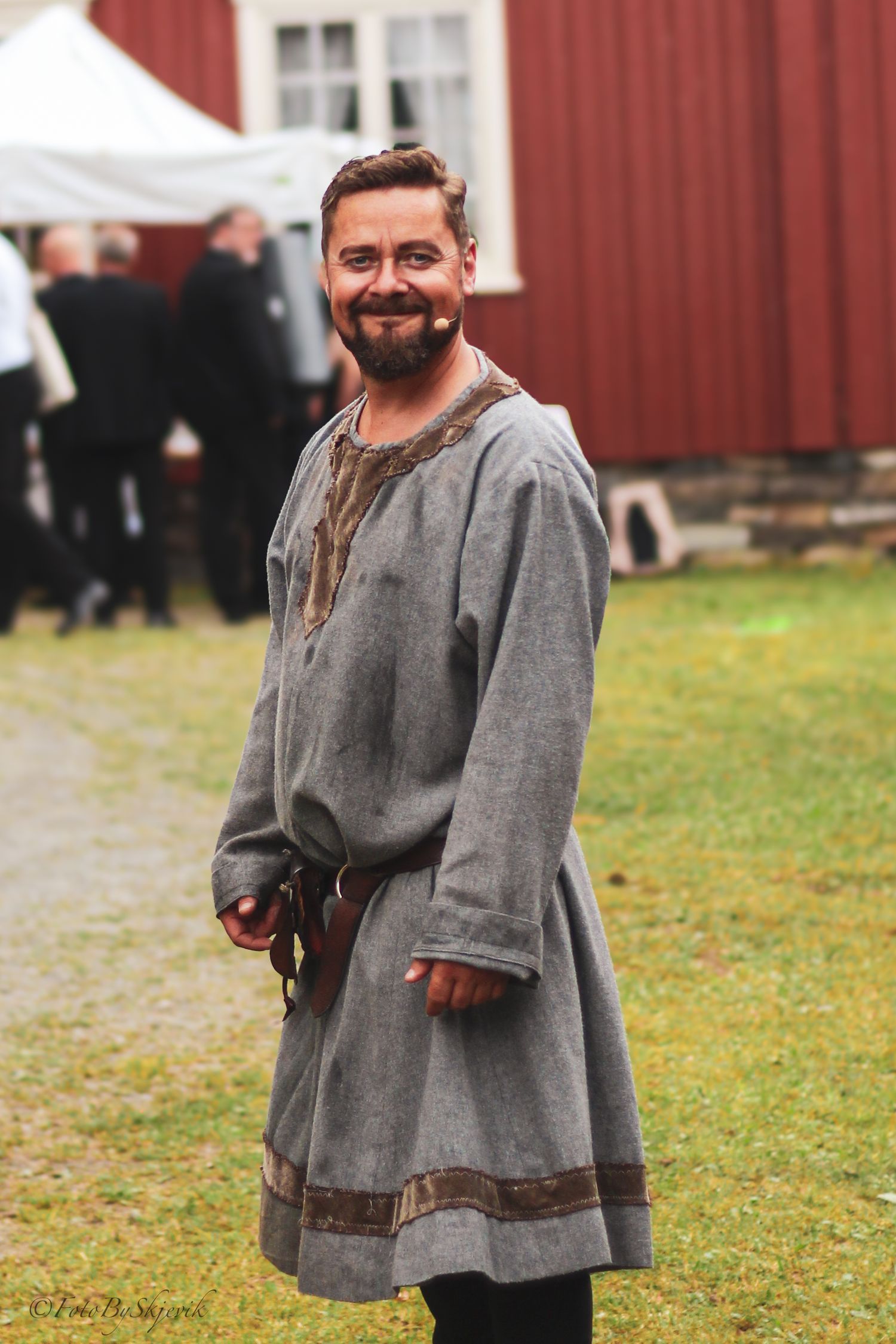 Person in historical grey tunic smiling near red barn buildings.