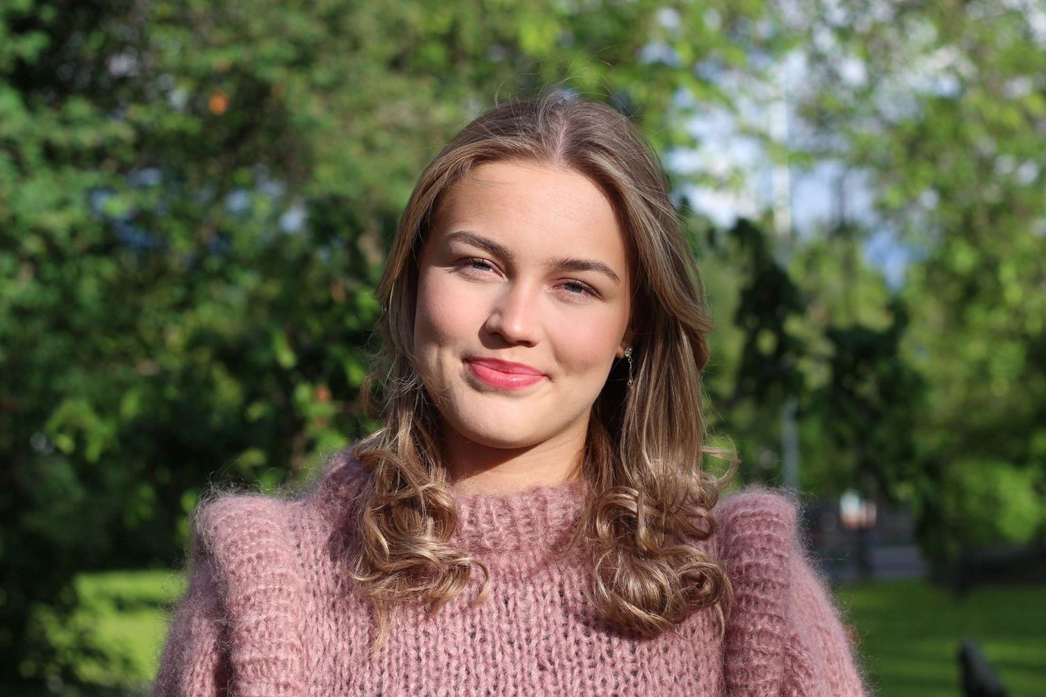 Woman wearing pink sweater smiles while posing in a garden setting.