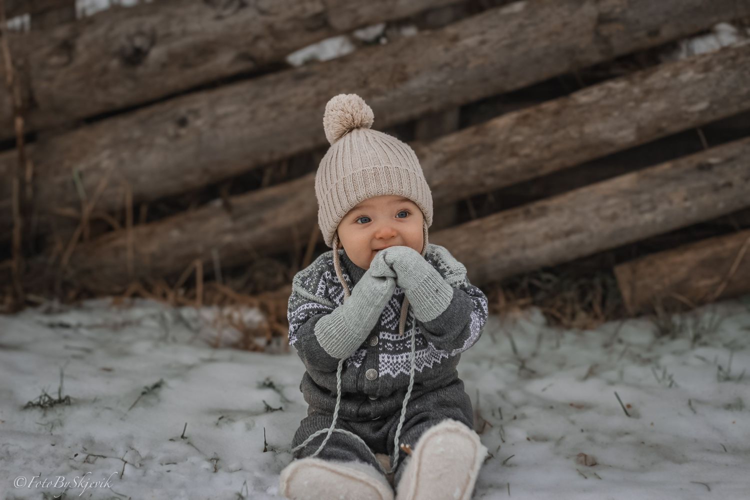 A cute baby in a knit hat and mittens sits in the snow near a rustic wooden fence in winter.