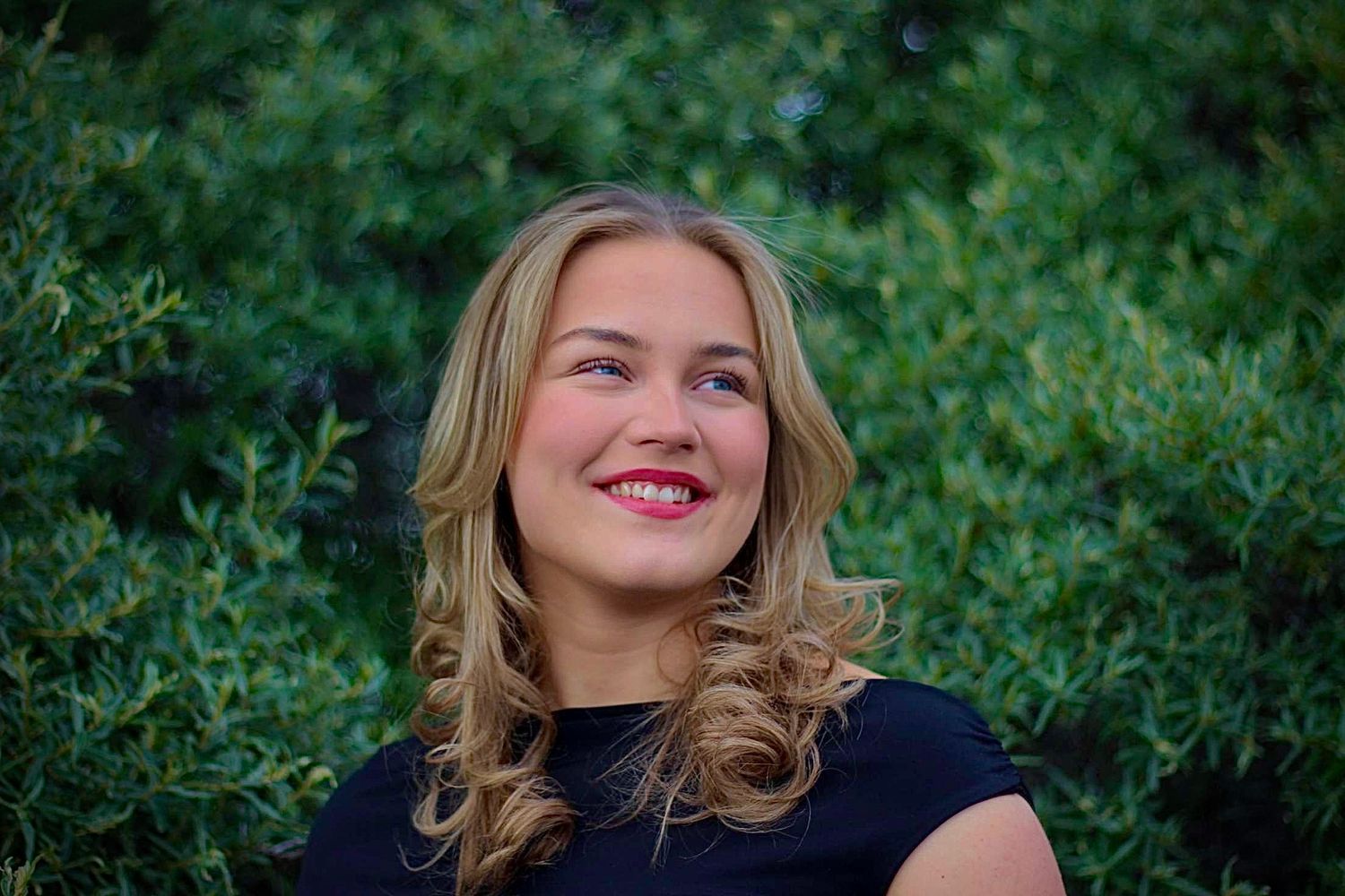 Woman with blonde hair poses outdoors against lush green foliage.