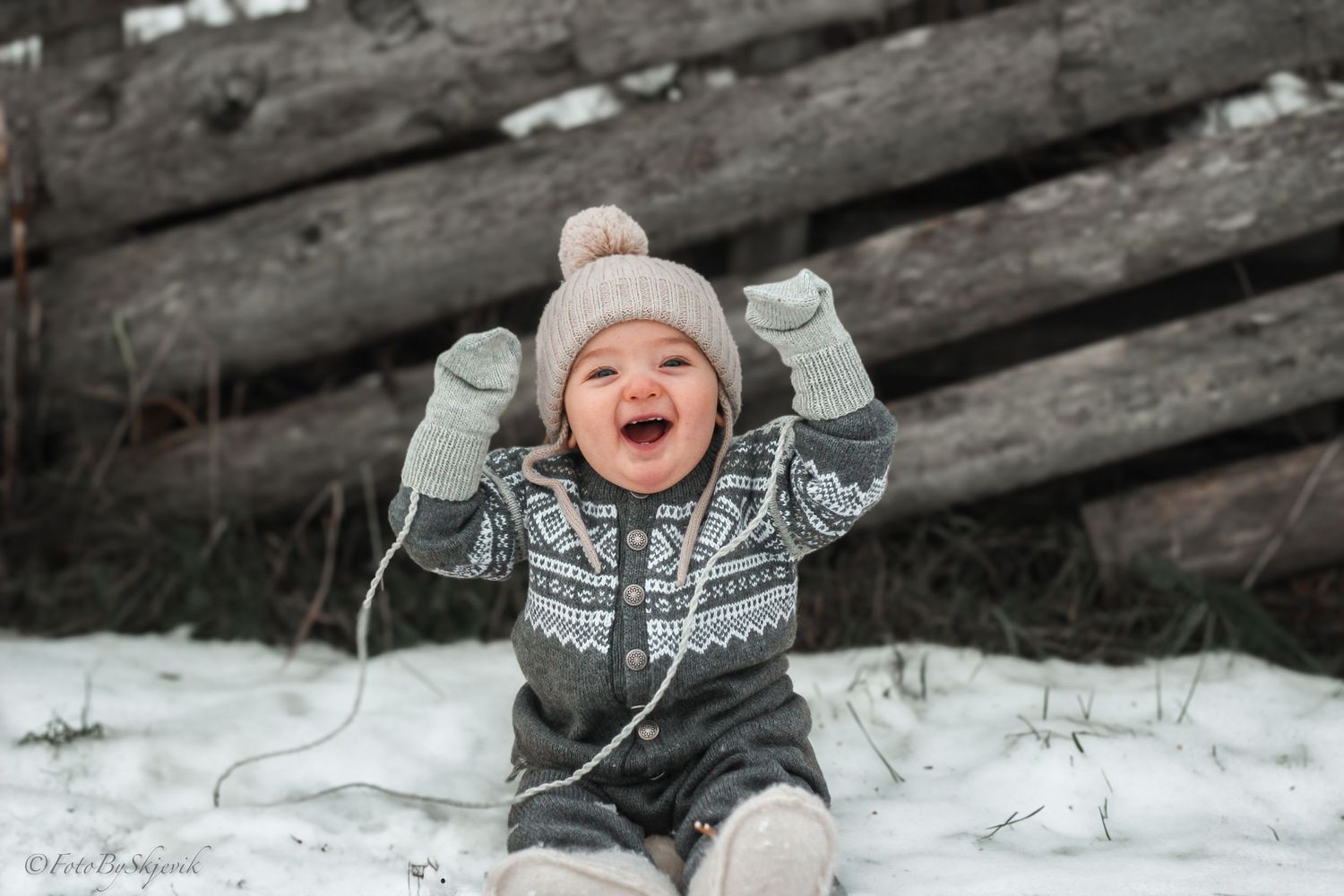 Happy baby in winter knit outfit and hat laughs joyfully while sitting in snow near rustic wooden fence.