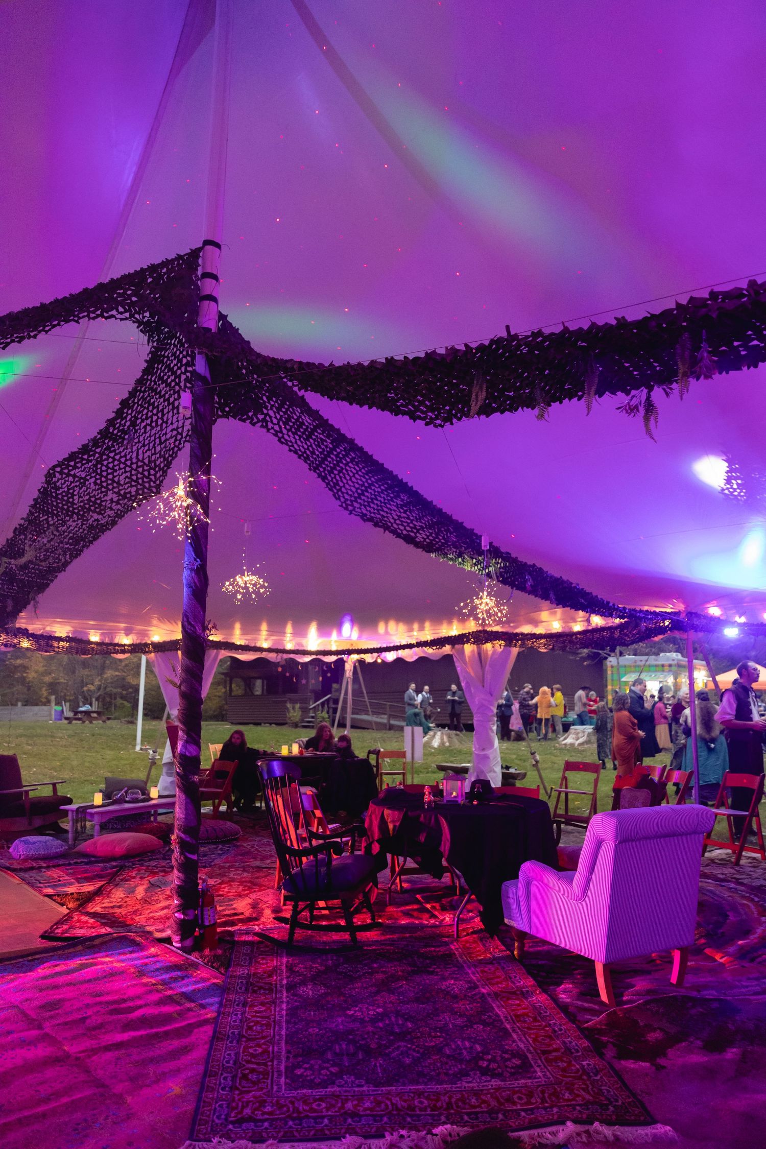 Elegant wedding reception tent glowing in purple light with chandeliers, lounge seating, and decorative draping.