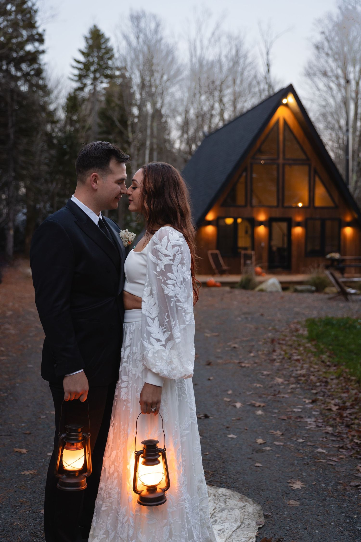 Bride and groom holding glowing lanterns outside a warmly lit A-frame cabin at dusk.