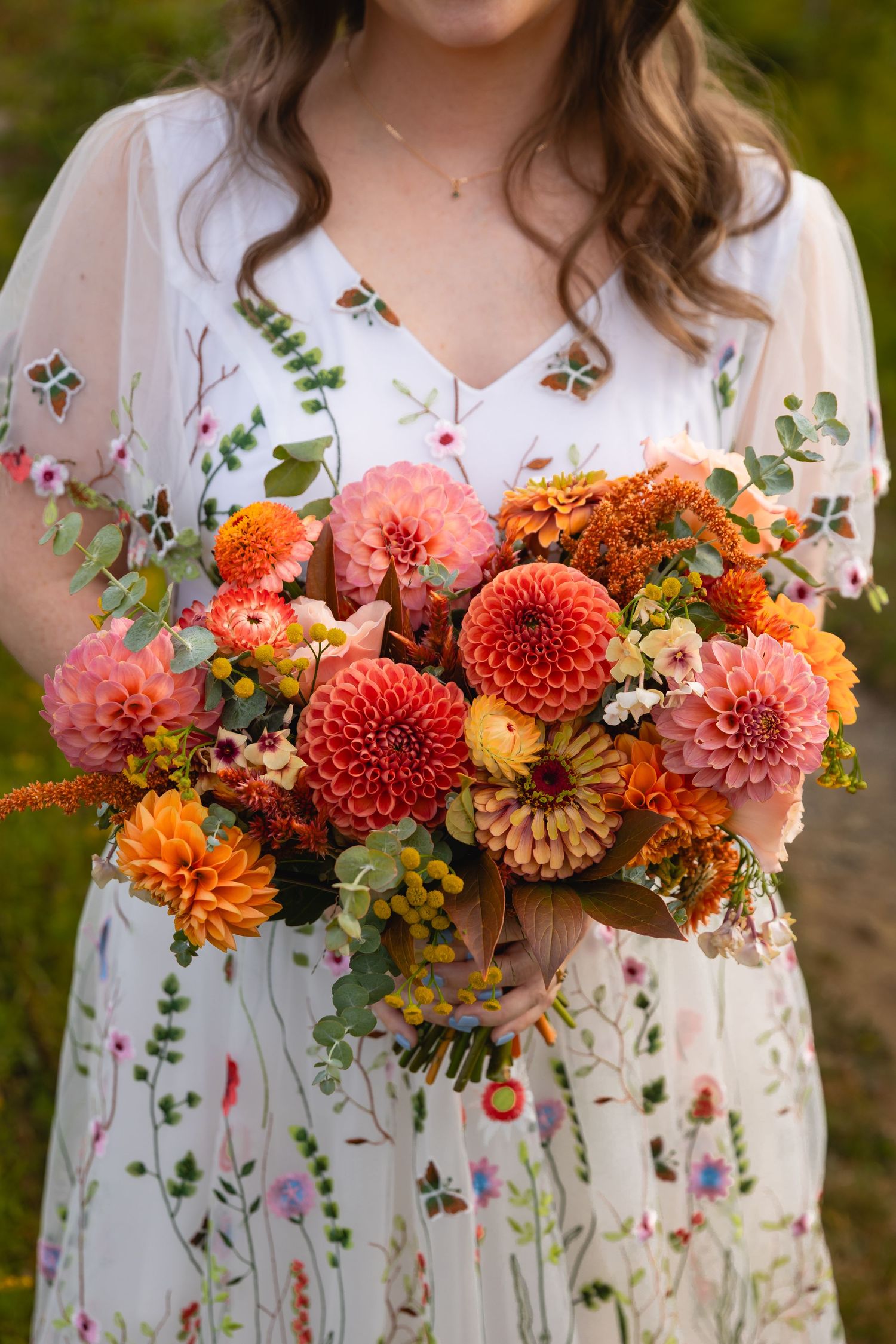 Beautiful autumn bridal bouquet with coral and orange dahlias held against a white floral dress with delicate embroidery.