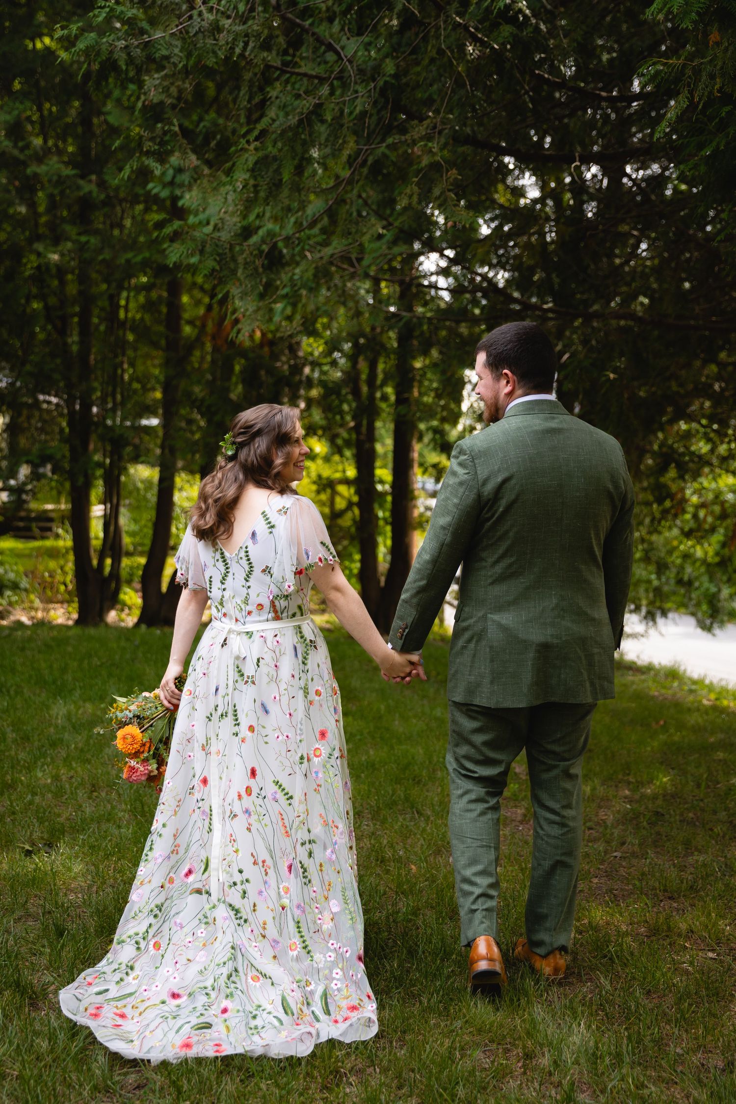 Couple in floral dress and green suit walking through a wooded park holding hands and picking up pumpkins.