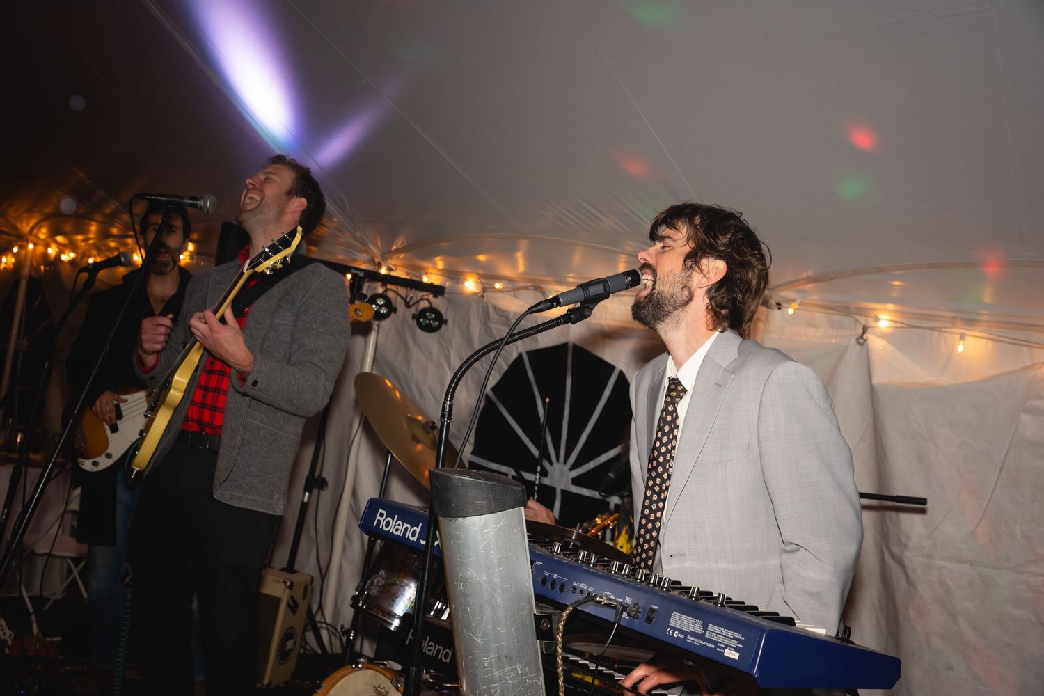 Musicians perform at a lively event under a tent with string lights and disco effects in the background.