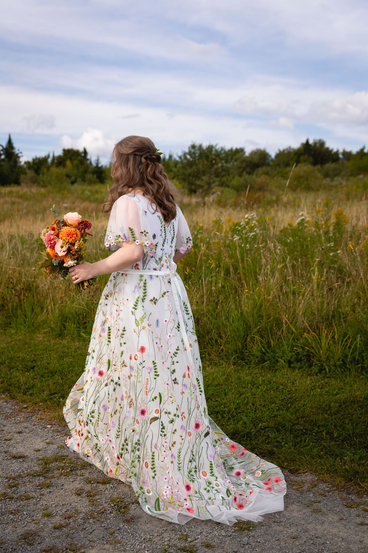A flowing white floral dress with embroidered wildflowers trails behind someone walking through a meadow at sunset.