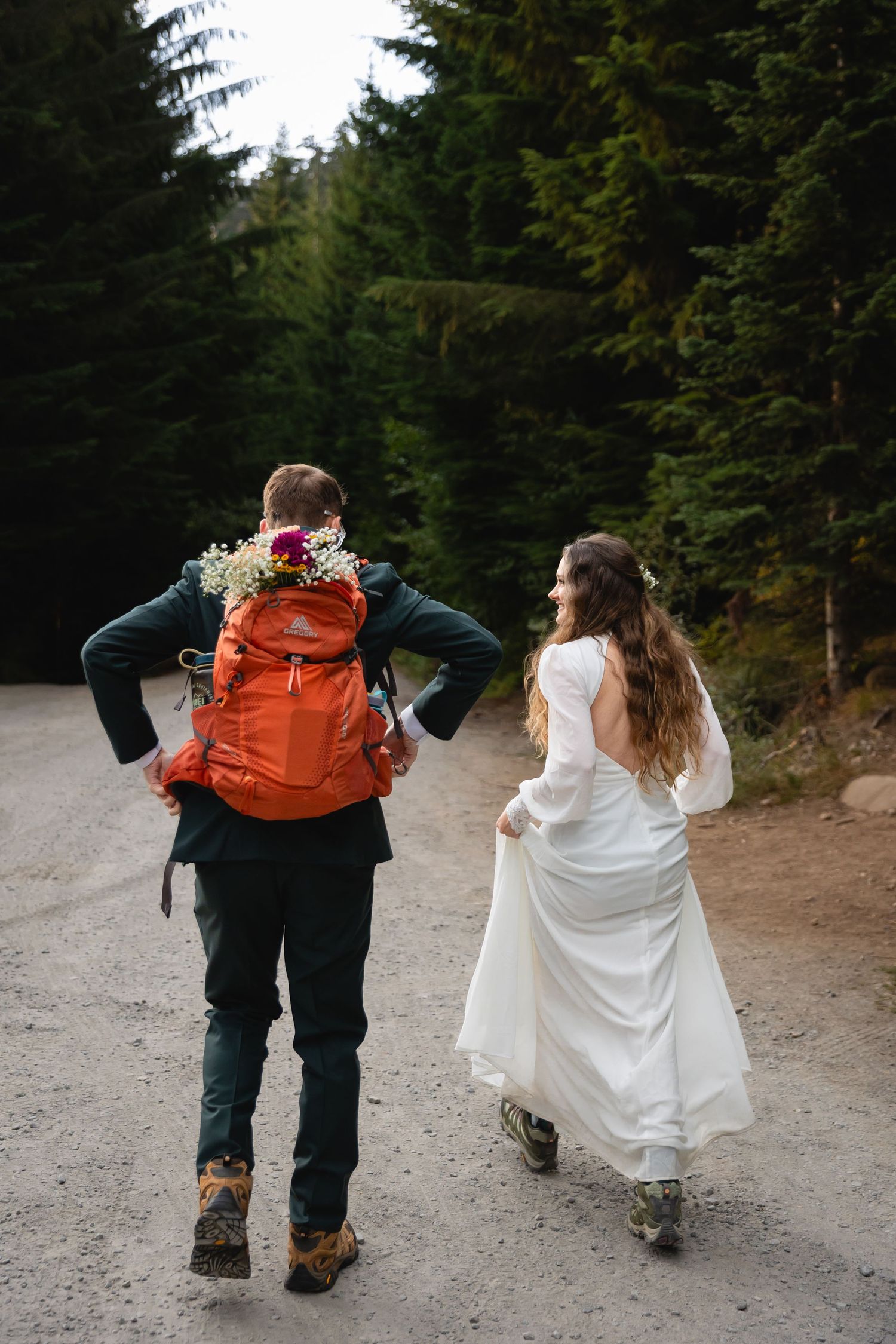 Two people in wedding attire walk along a mountain trail carrying backpacks and hiking gear.