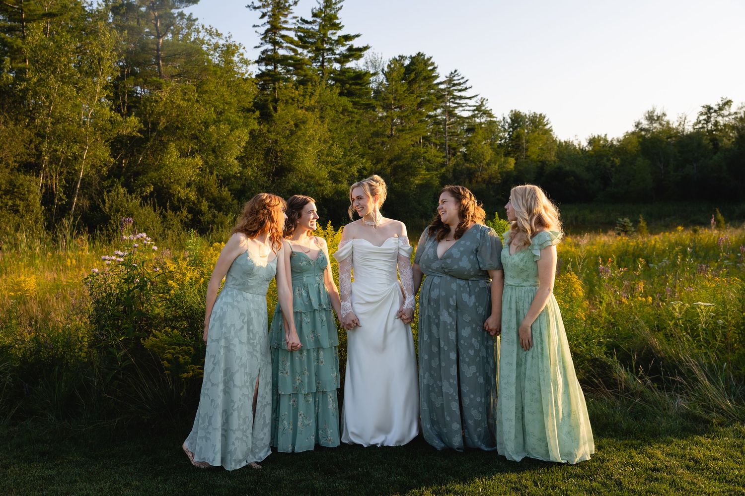 Group in soft blue dresses and white dress standing together in a golden sunlit meadow at sunset.