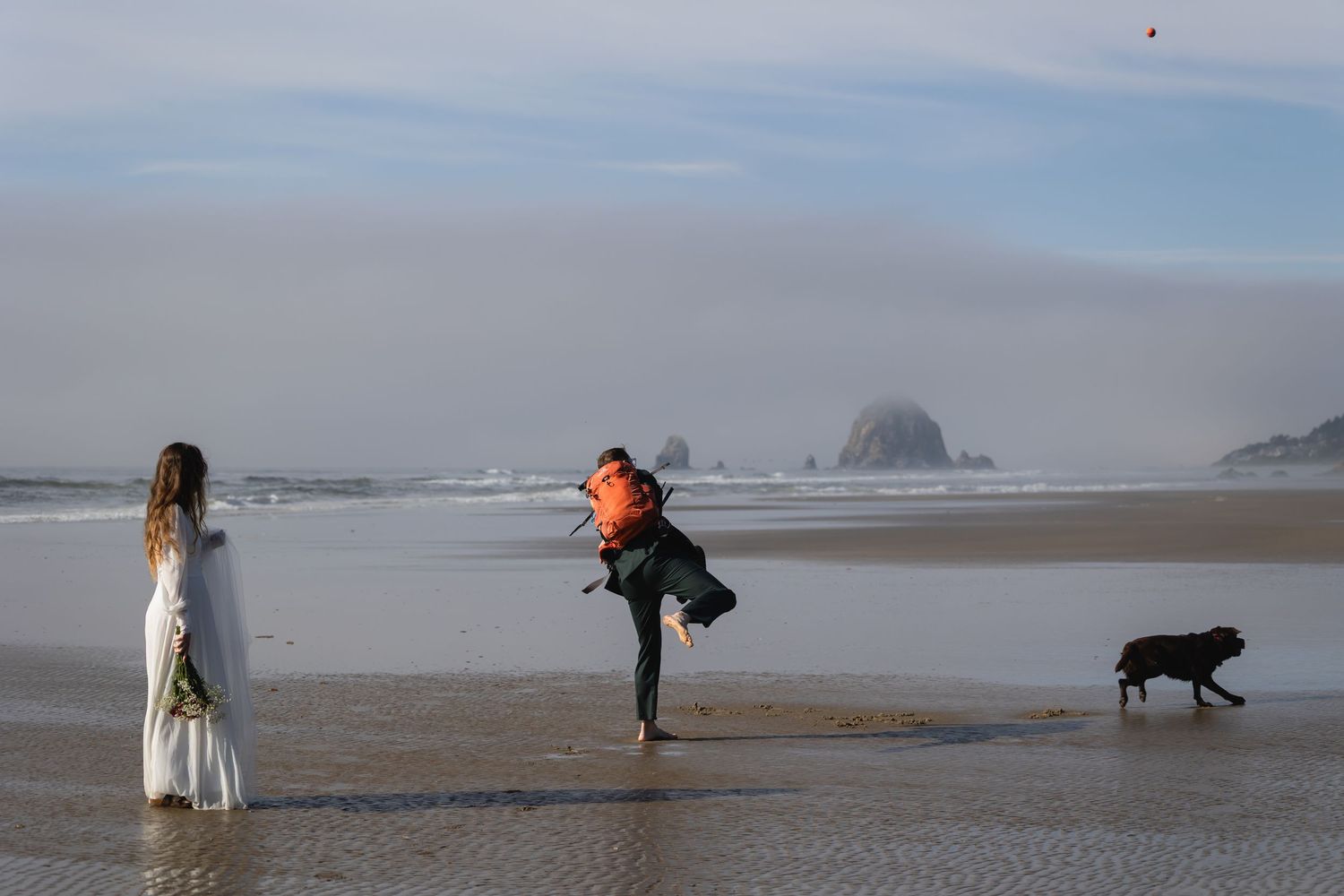 People and a dog walk along a misty beach with rocky sea stacks visible in the distance.