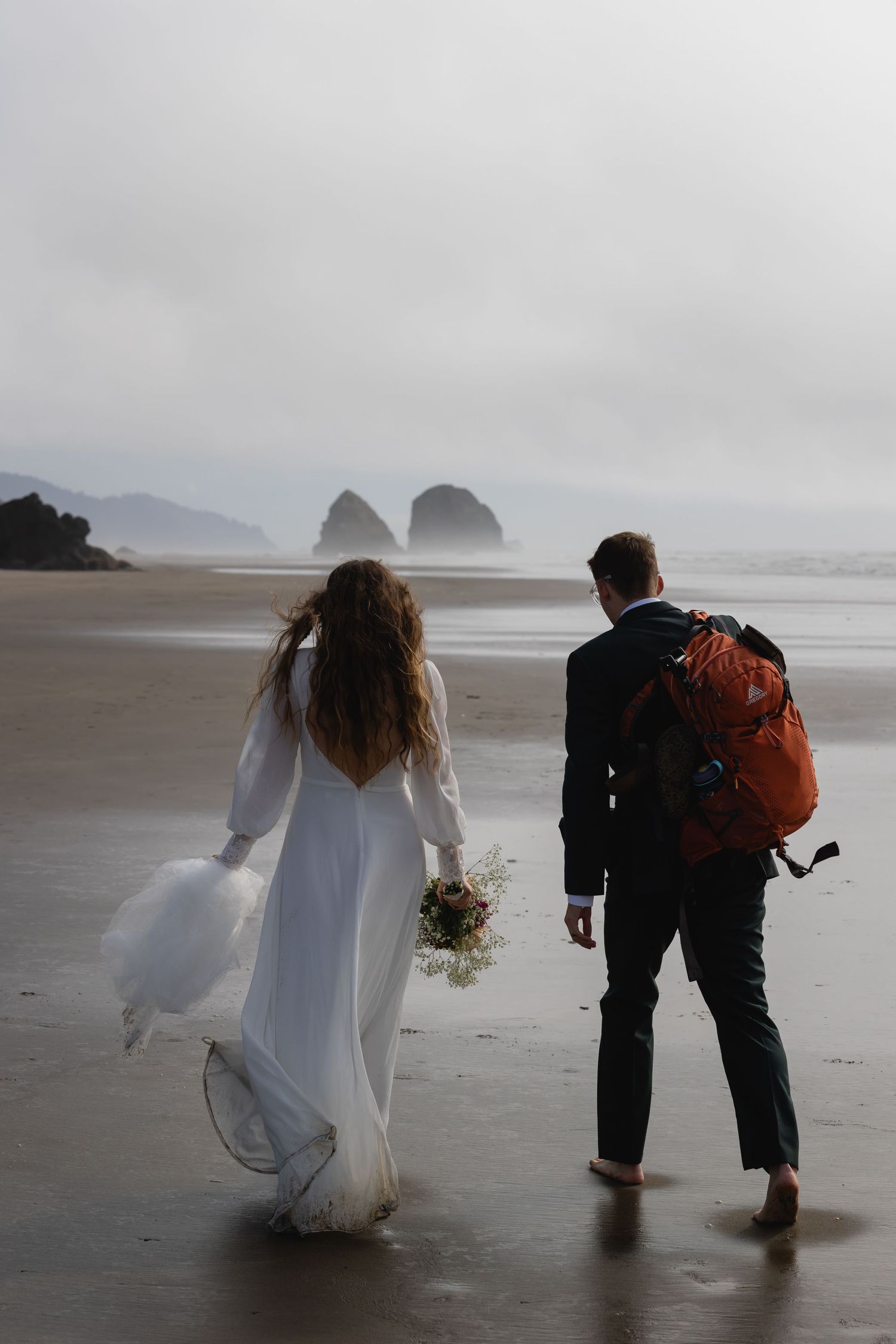 A couple walks barefoot on a misty beach with dramatic rock formations in the background at Cannon Beach, Oregon.