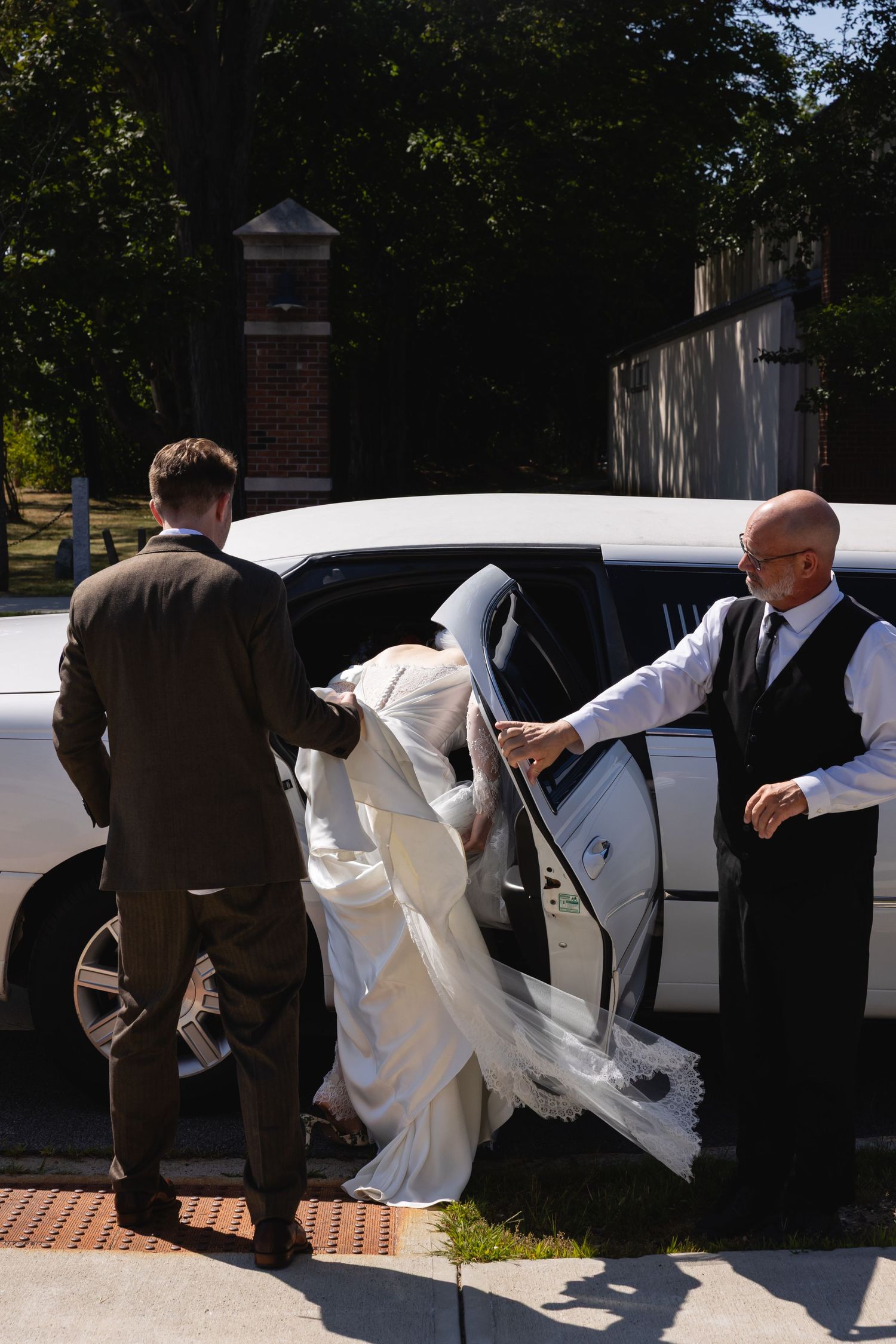 A white wedding dress flowing into a luxury car while attendants assist outside on a sunny day.