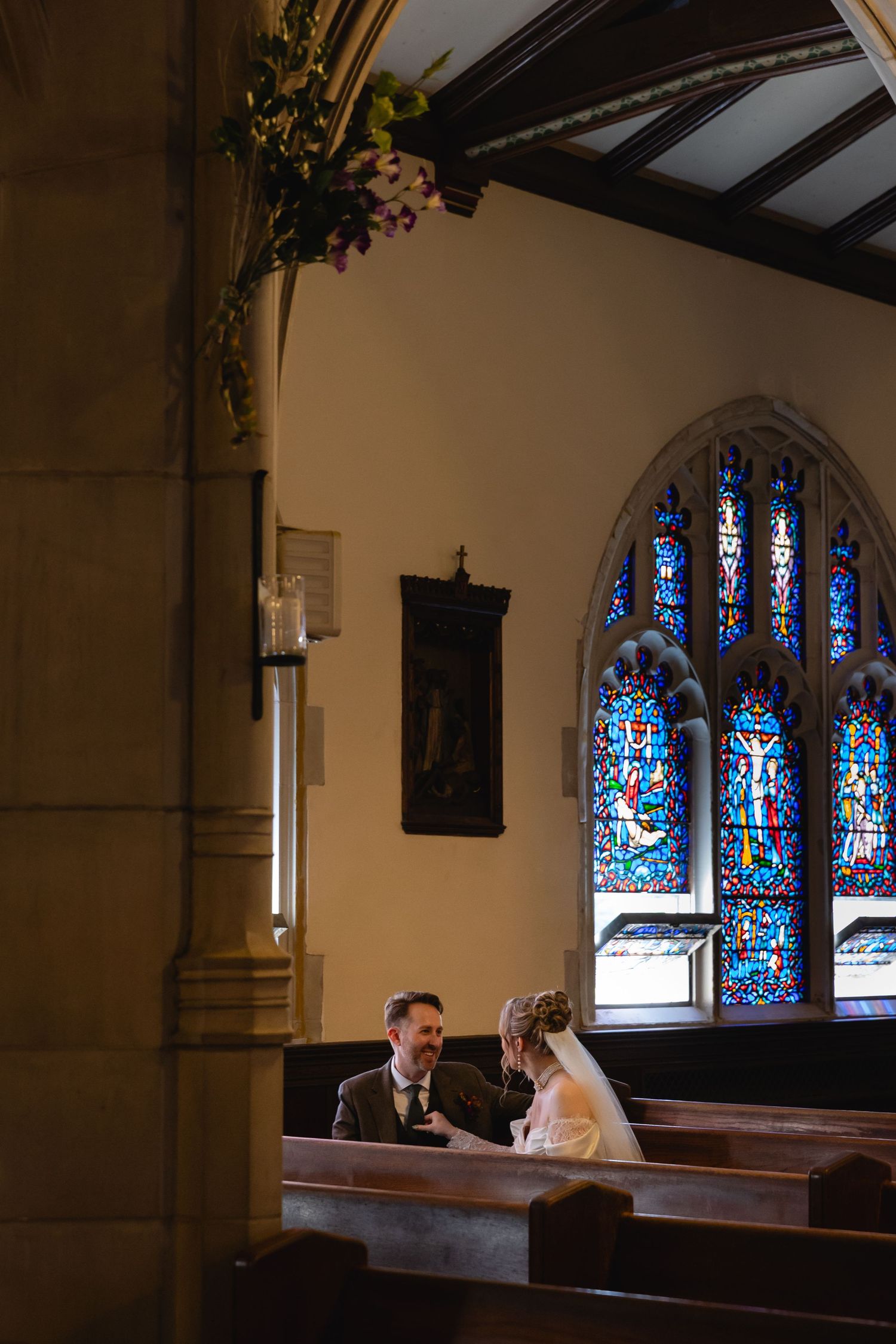 Couple sitting in church pews illuminated by colorful stained glass windows during an intimate wedding ceremony.