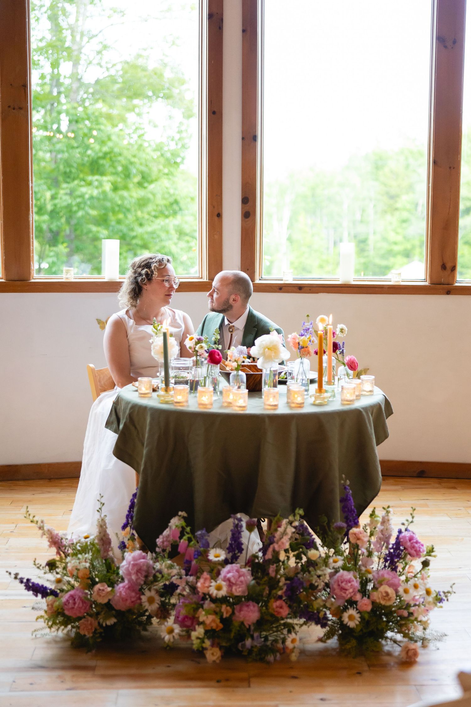 A romantic sweetheart table adorned with pink and purple floral arrangements at a wedding reception venue with natural lighting.